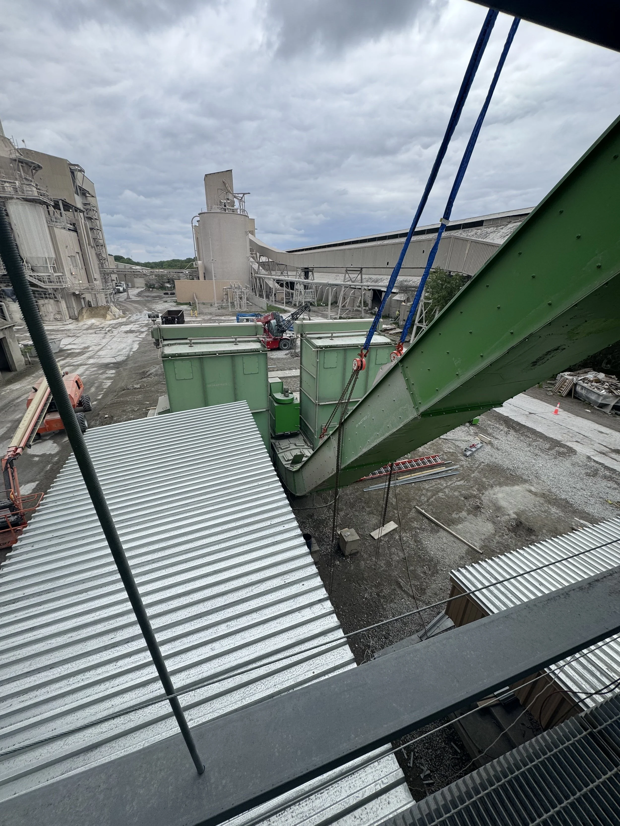 View of an industrial construction site with green metal structures, large storage tanks, and machinery under a cloudy sky.