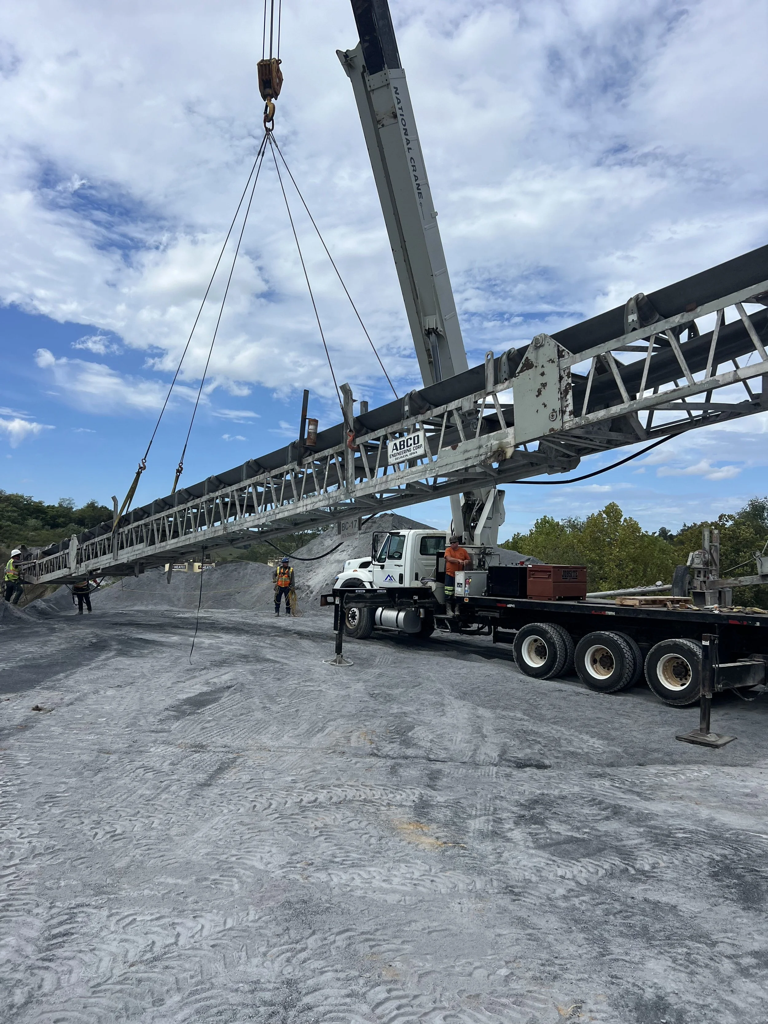 Construction workers operating a crane and a conveyor belt at a construction site under a partly cloudy sky.