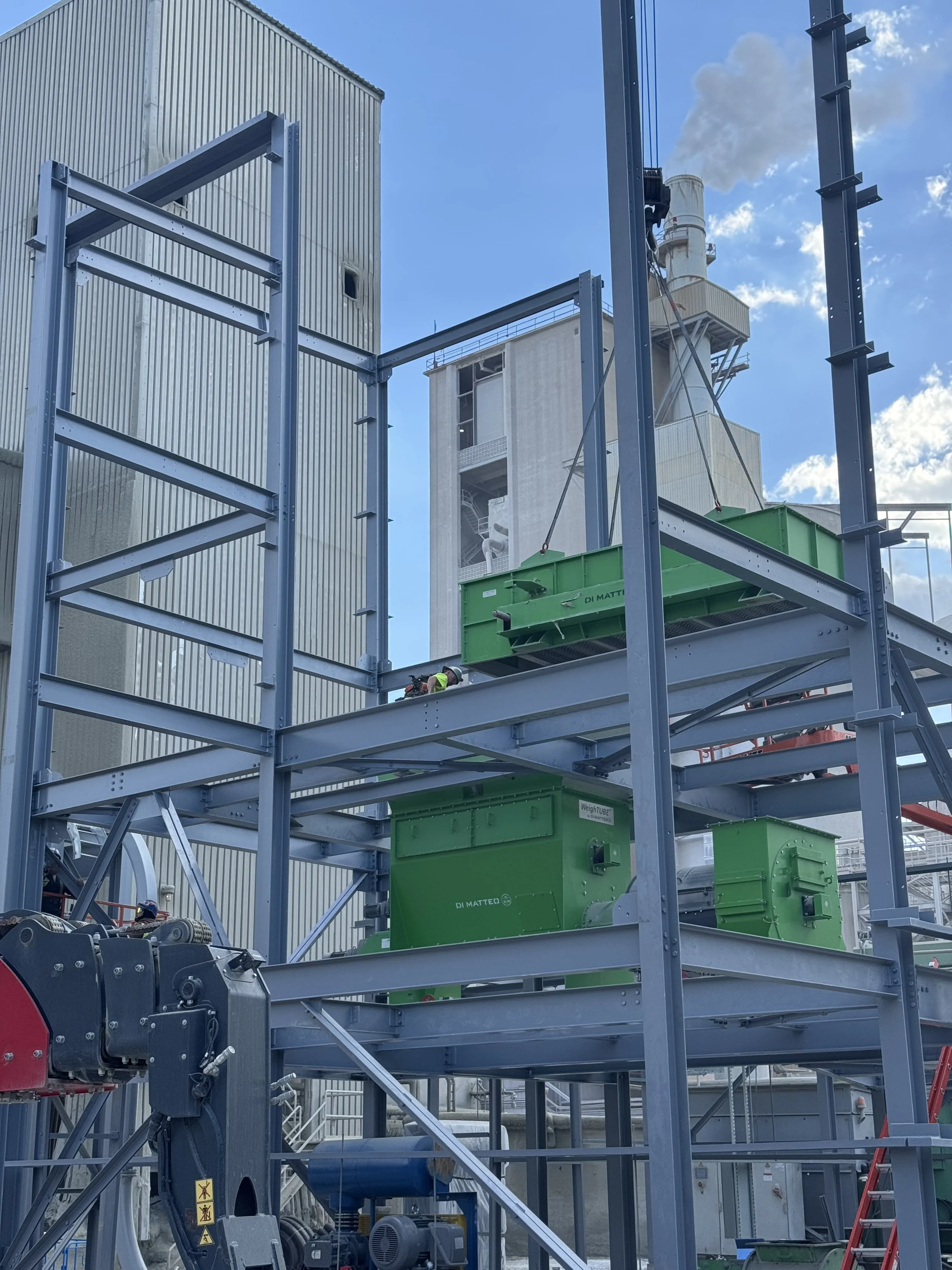 Construction site with metal framework, large green machinery labeled 'DI MATTEO', industrial buildings in the background, and a clear sky with some clouds.