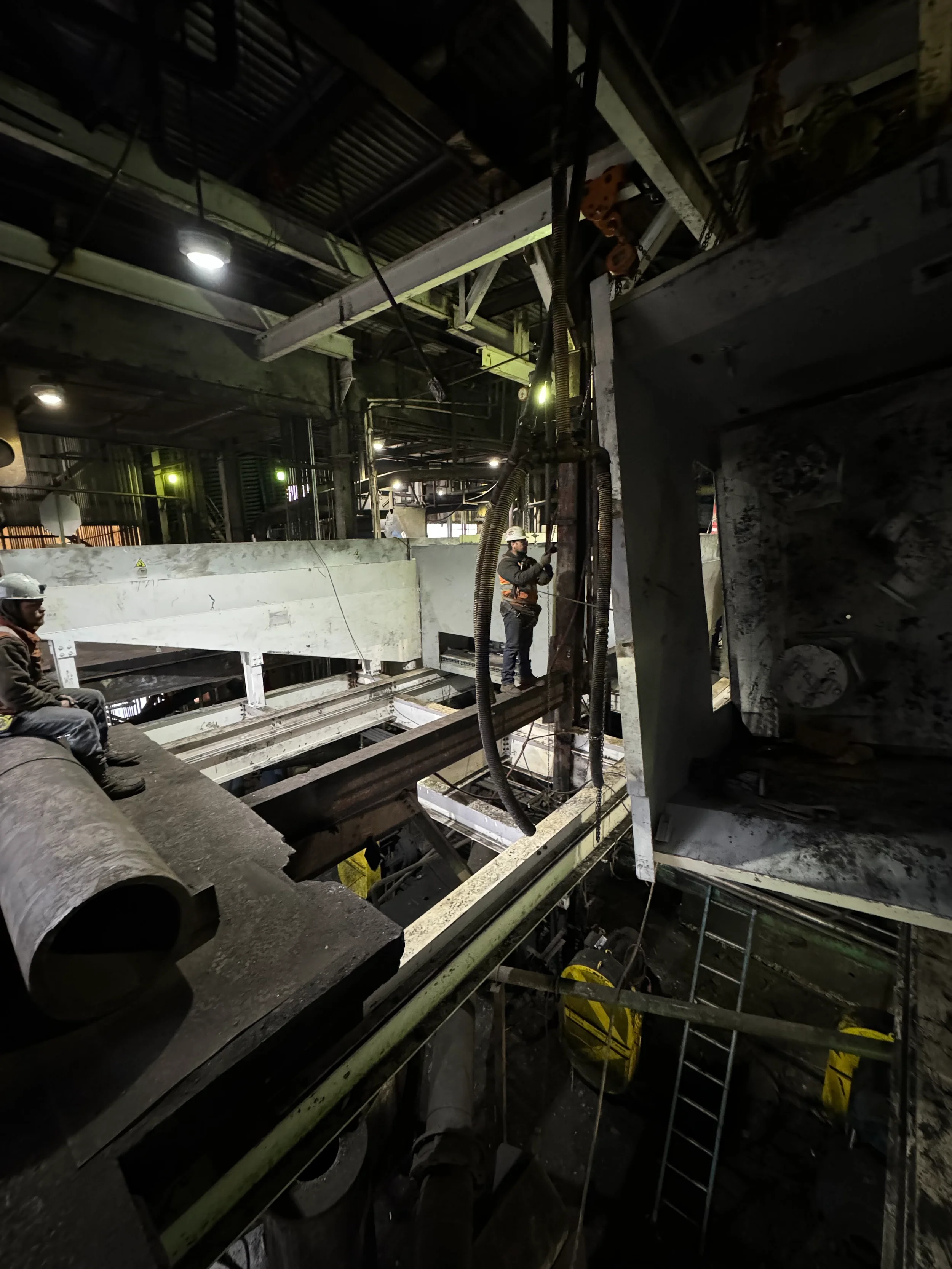 Workers in hard hats and safety gear working inside an industrial facility with metal beams, pipes, and machinery, illuminated by overhead lights.