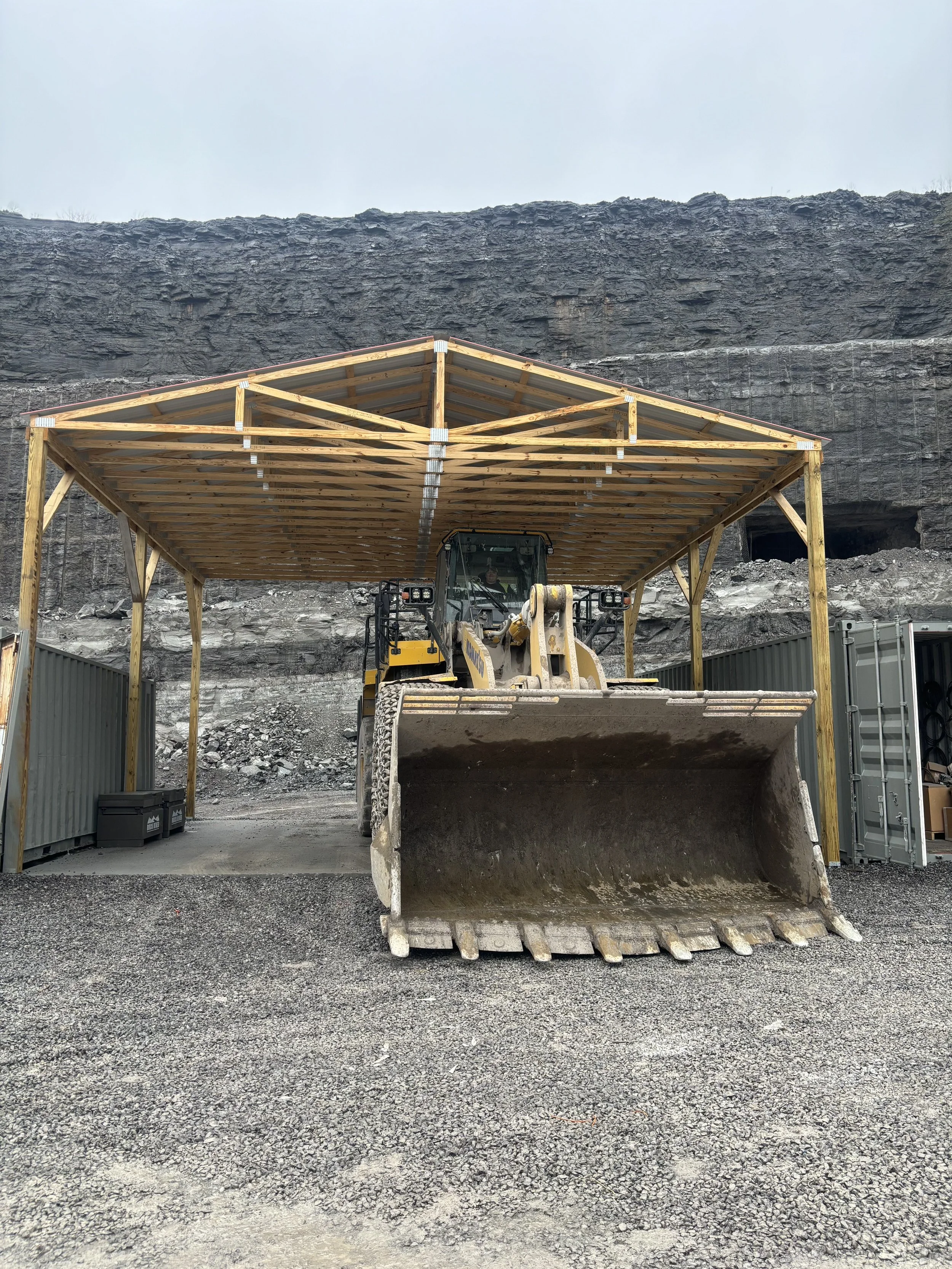 Construction site with a front loader under a wooden shelter, rocky mountain in the background, and gravel ground.