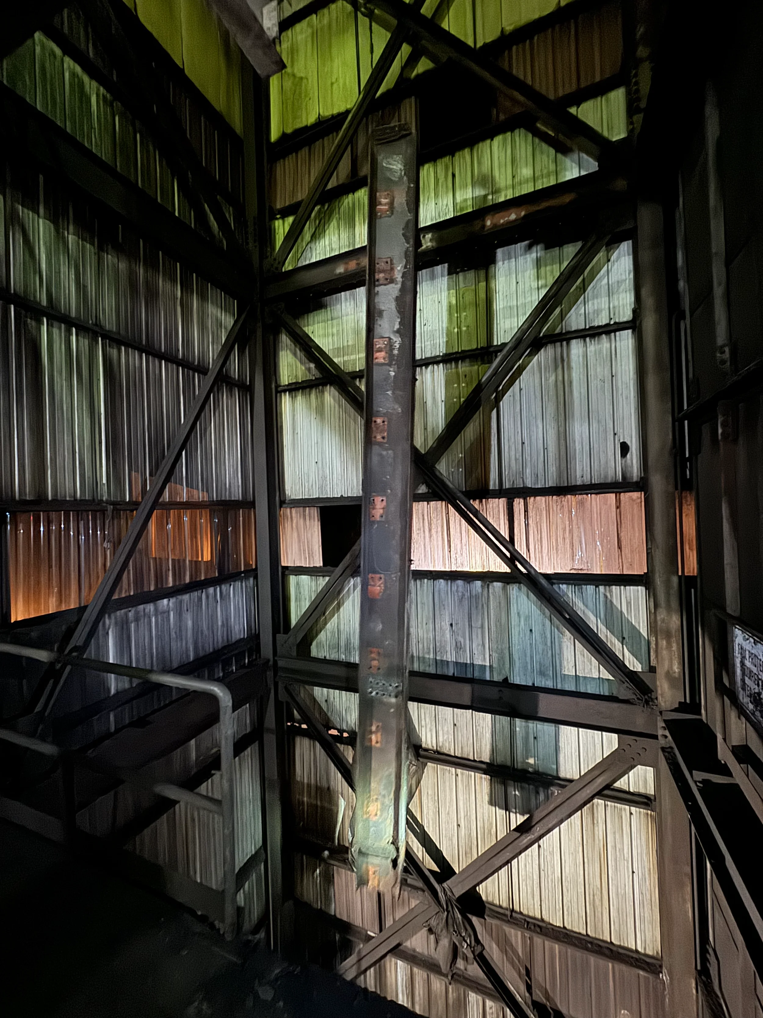 Interior view of a metal staircase with a multi-colored corrugated metal wall in the background, featuring vertical panels in shades of green, beige, and orange.