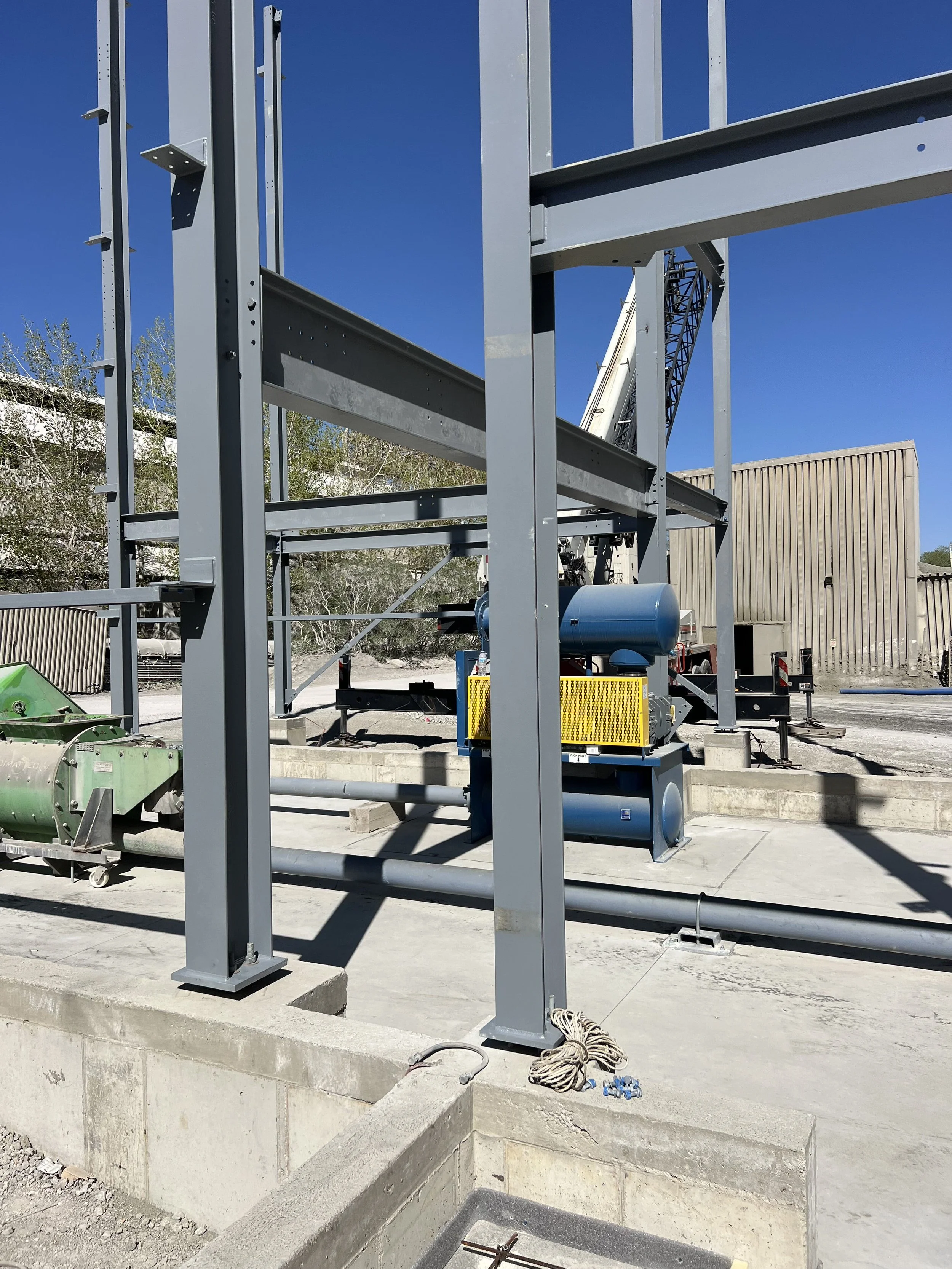 Construction site with metal framework for a building, a crane in the background, and construction equipment on the ground under a clear blue sky.
