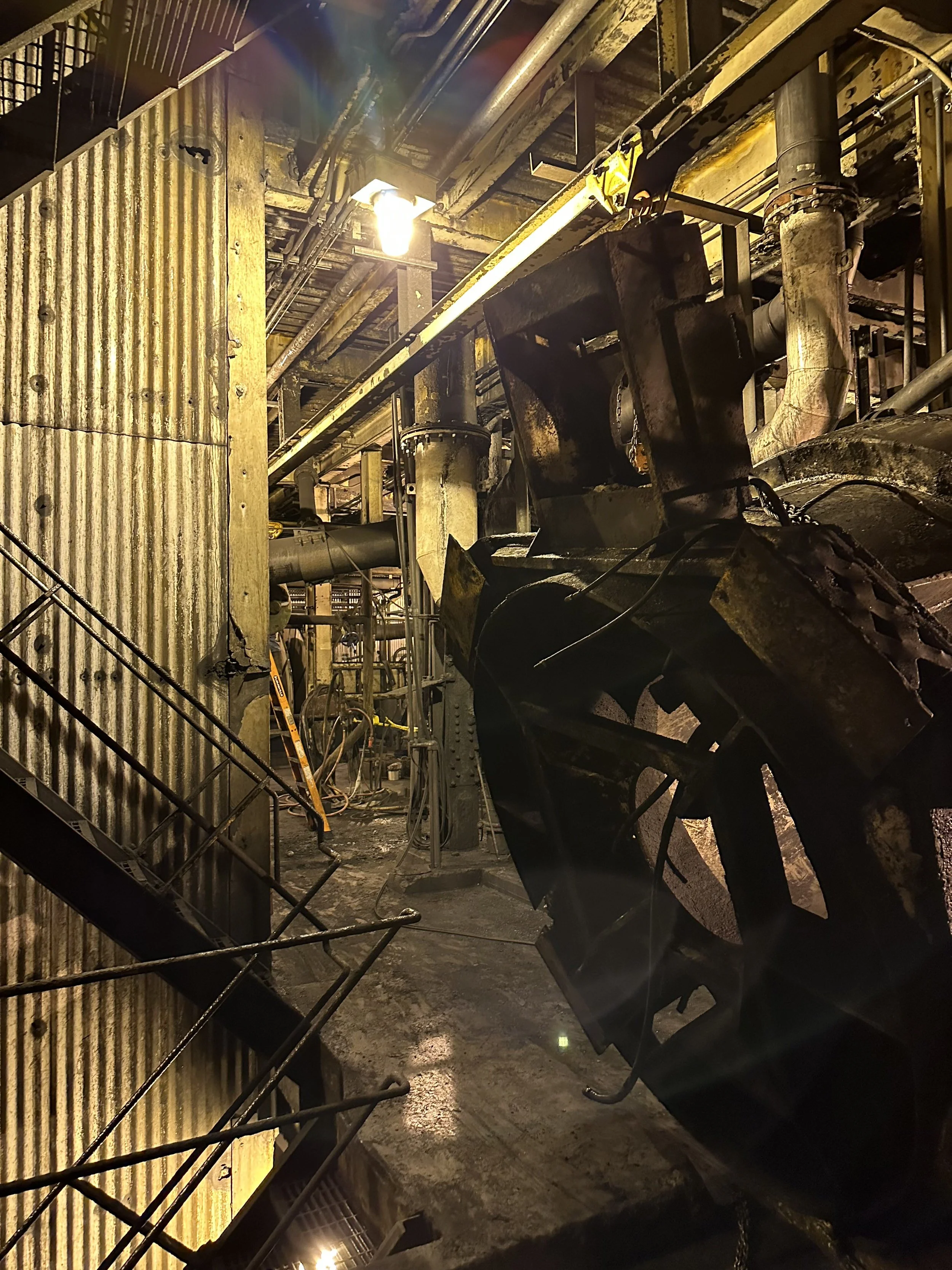 The image shows an industrial setting with metal pipes, machinery, and a corrugated tin wall. The lighting is warm, and there is a ladder and safety chains visible, indicating construction or maintenance work.