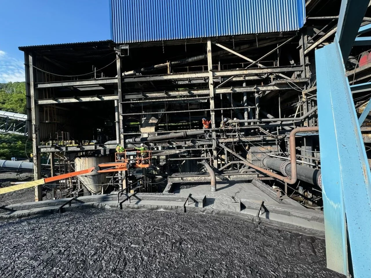 Industrial facility with metal framework, pipes, and workers wearing safety gear working on equipment, with a blackened area in the foreground and a blue building at the top.