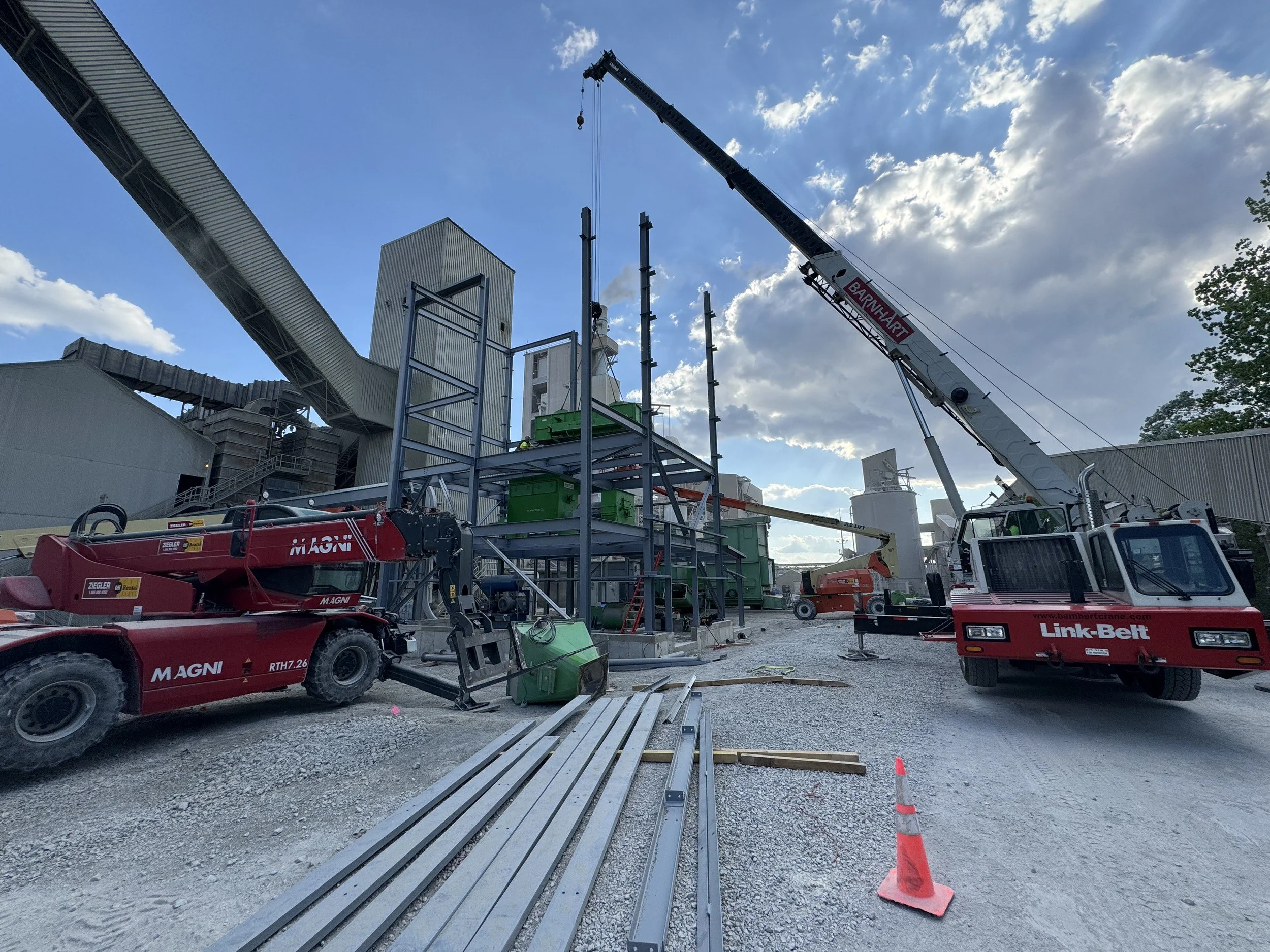 Construction site with cranes and equipment, metal beams on the ground, and a building structure under construction against a partly cloudy sky.