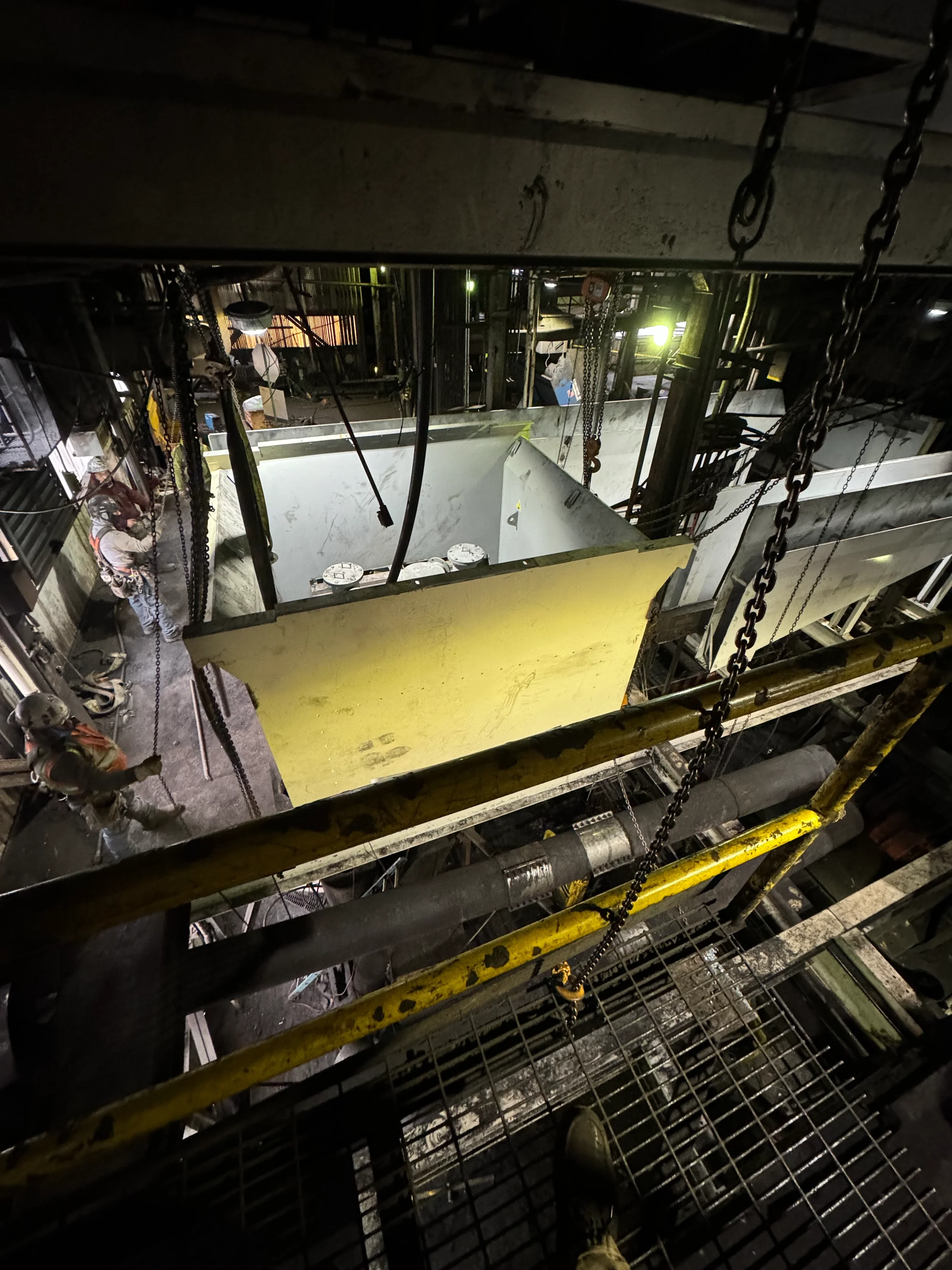 View of an industrial or construction site with metal scaffolding and chains, illuminated by a yellowish light. A worker is present on the left side, wearing protective gear.