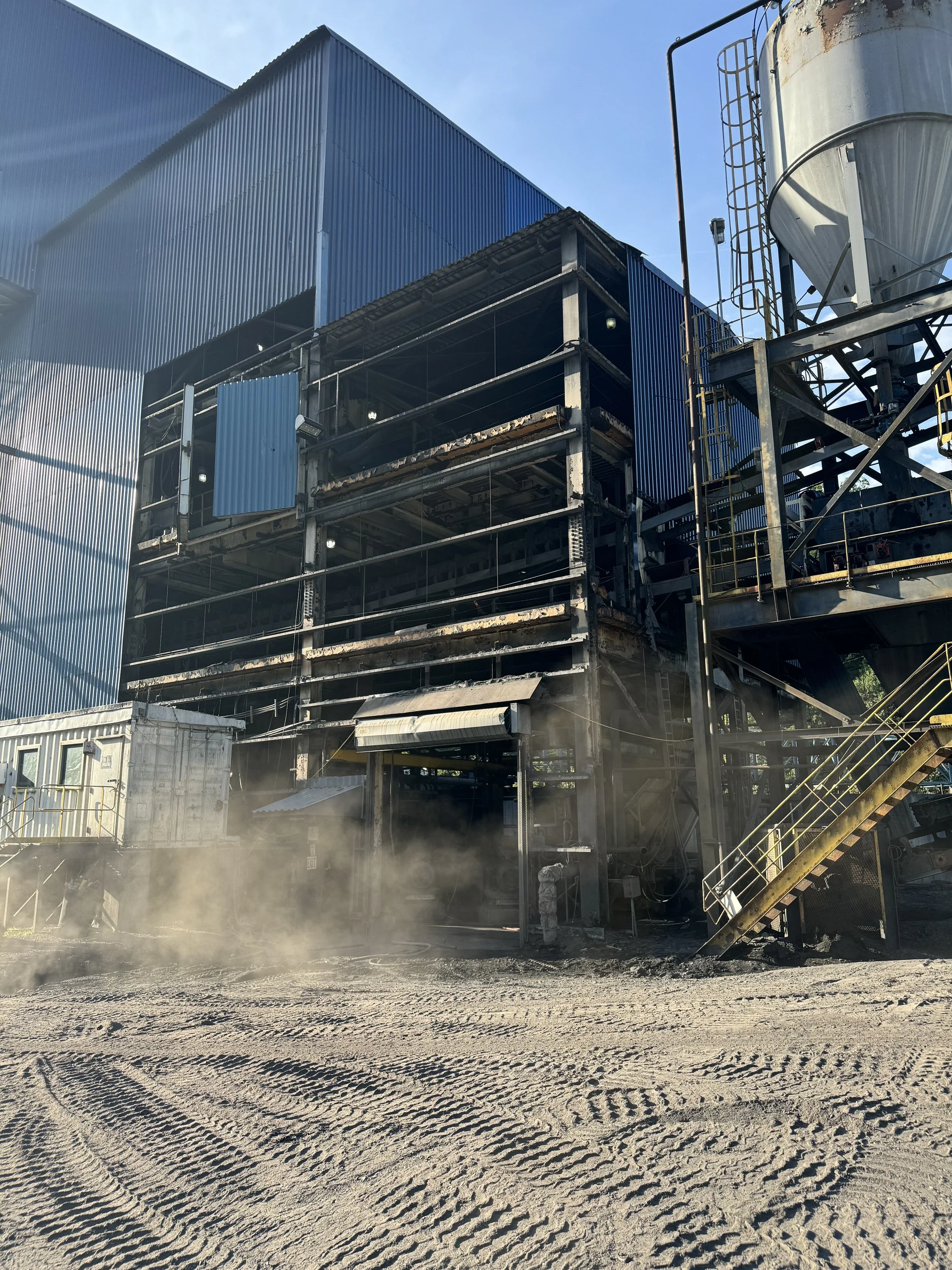 A construction site with a large industrial building under construction, metal scaffolding, and a large silo, with dirt ground and dust in the air.
