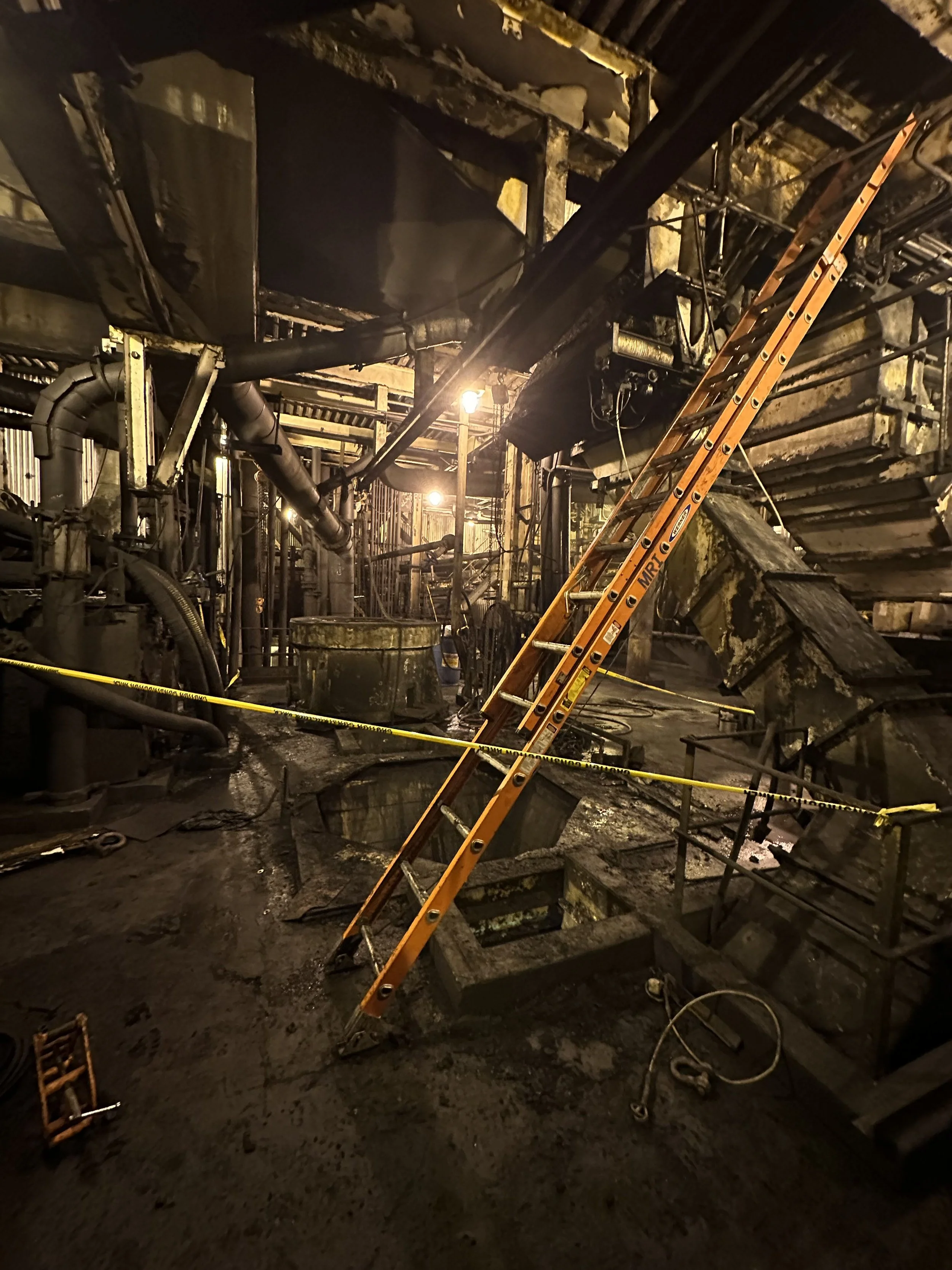 Interior of a building with extensive fire damage, showing charred and melted structural elements, soot-covered surfaces, and an orange ladder leaning against the collapsed ceiling.
