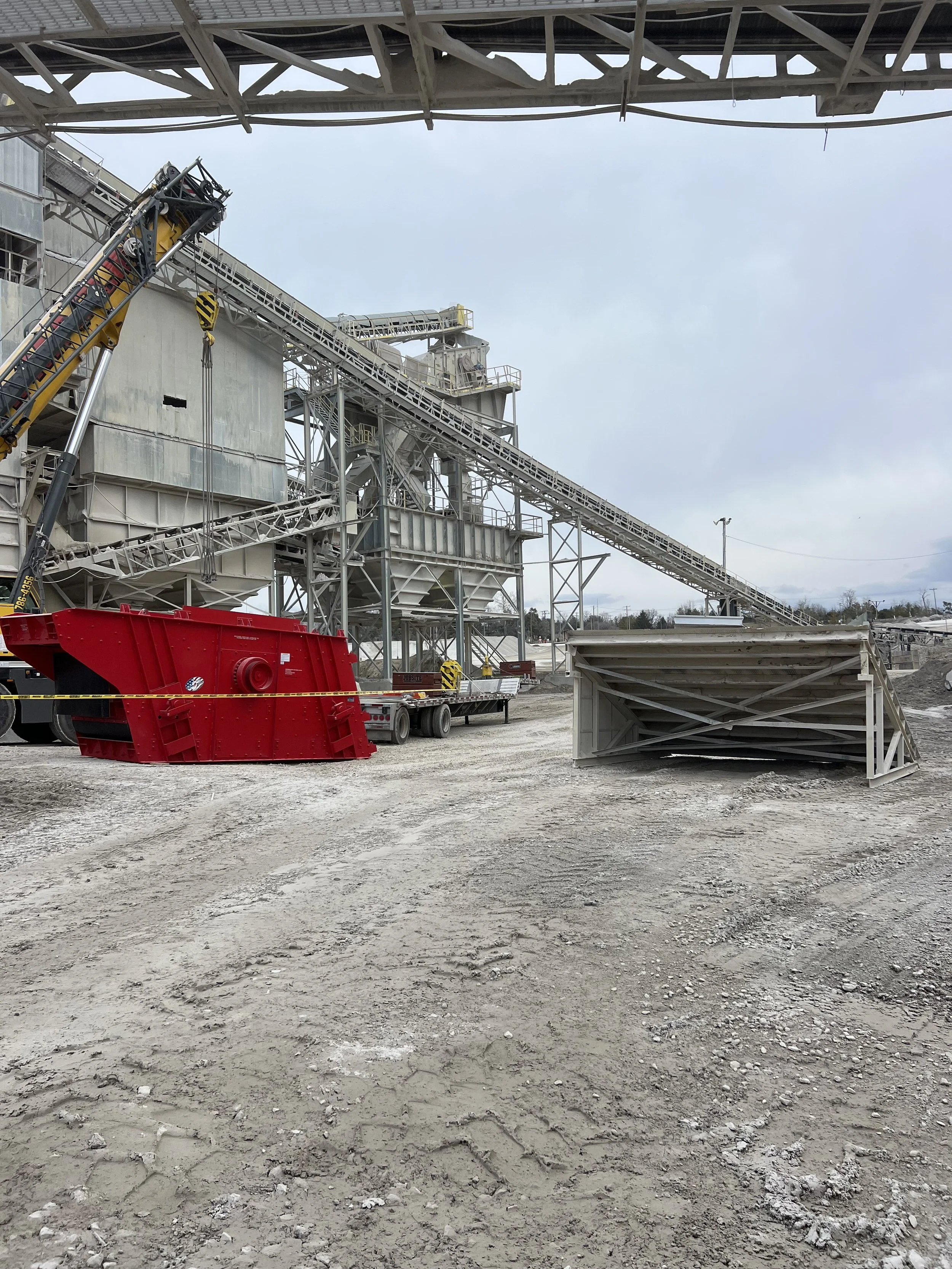 Construction site with industrial machinery, metal structures, and a gravel ground. Overcast sky.