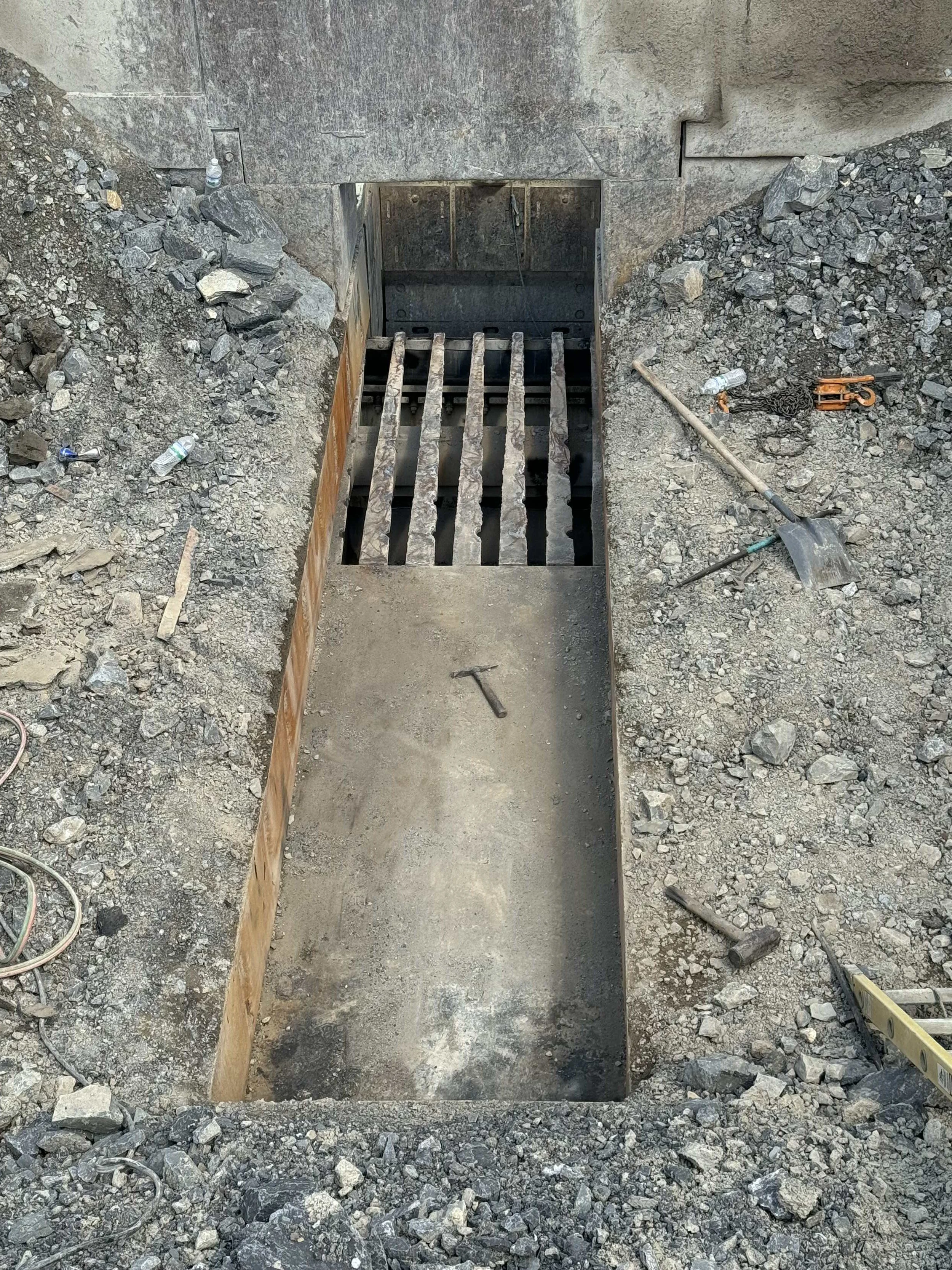 Construction site showing a trench with metal grating, surrounded by dirt, rocks, and construction tools, with a wooden barrier on the left side.
