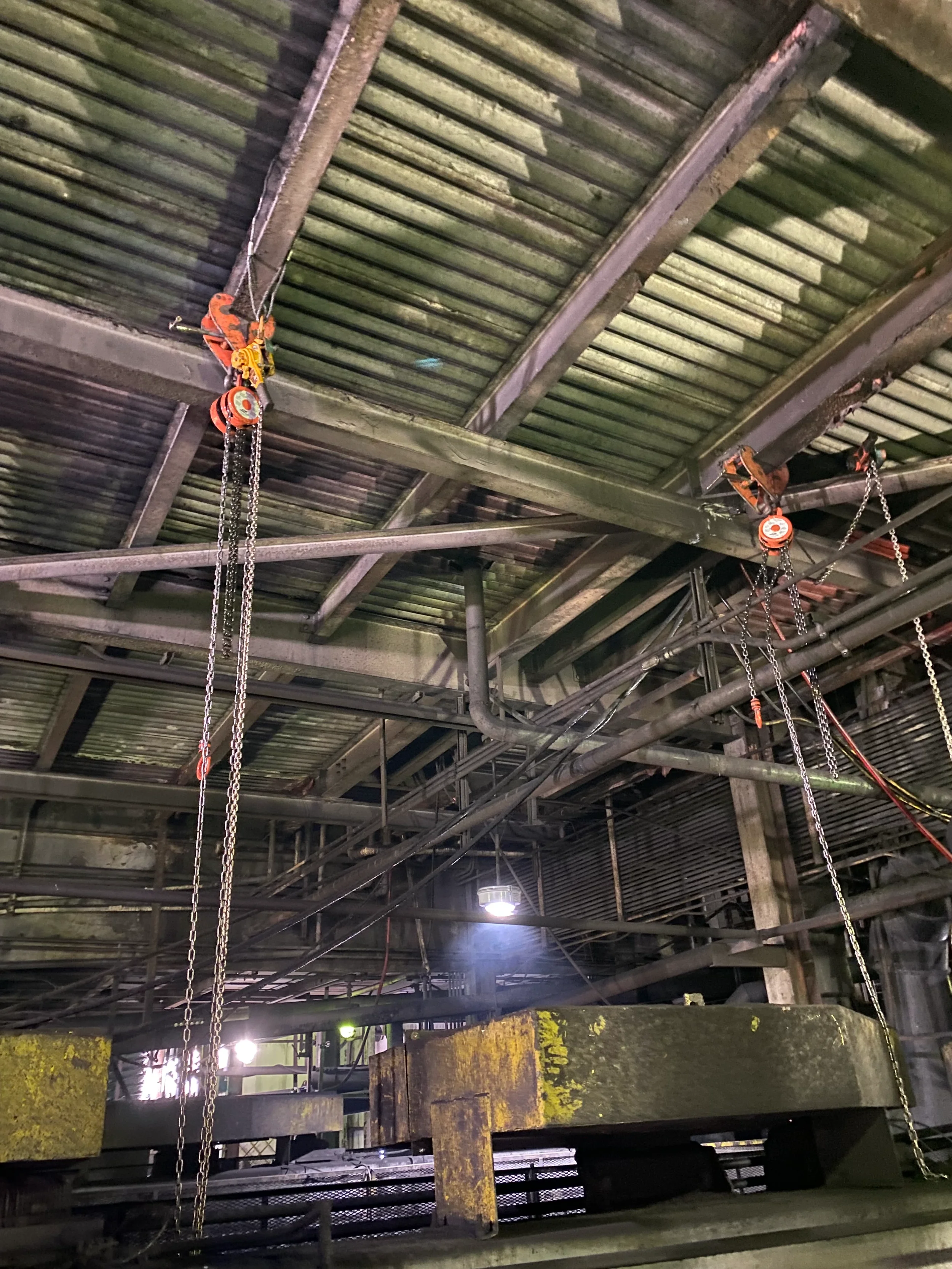 Industrial warehouse ceiling with metal beams, chains, and hanging equipment, with dim lighting.