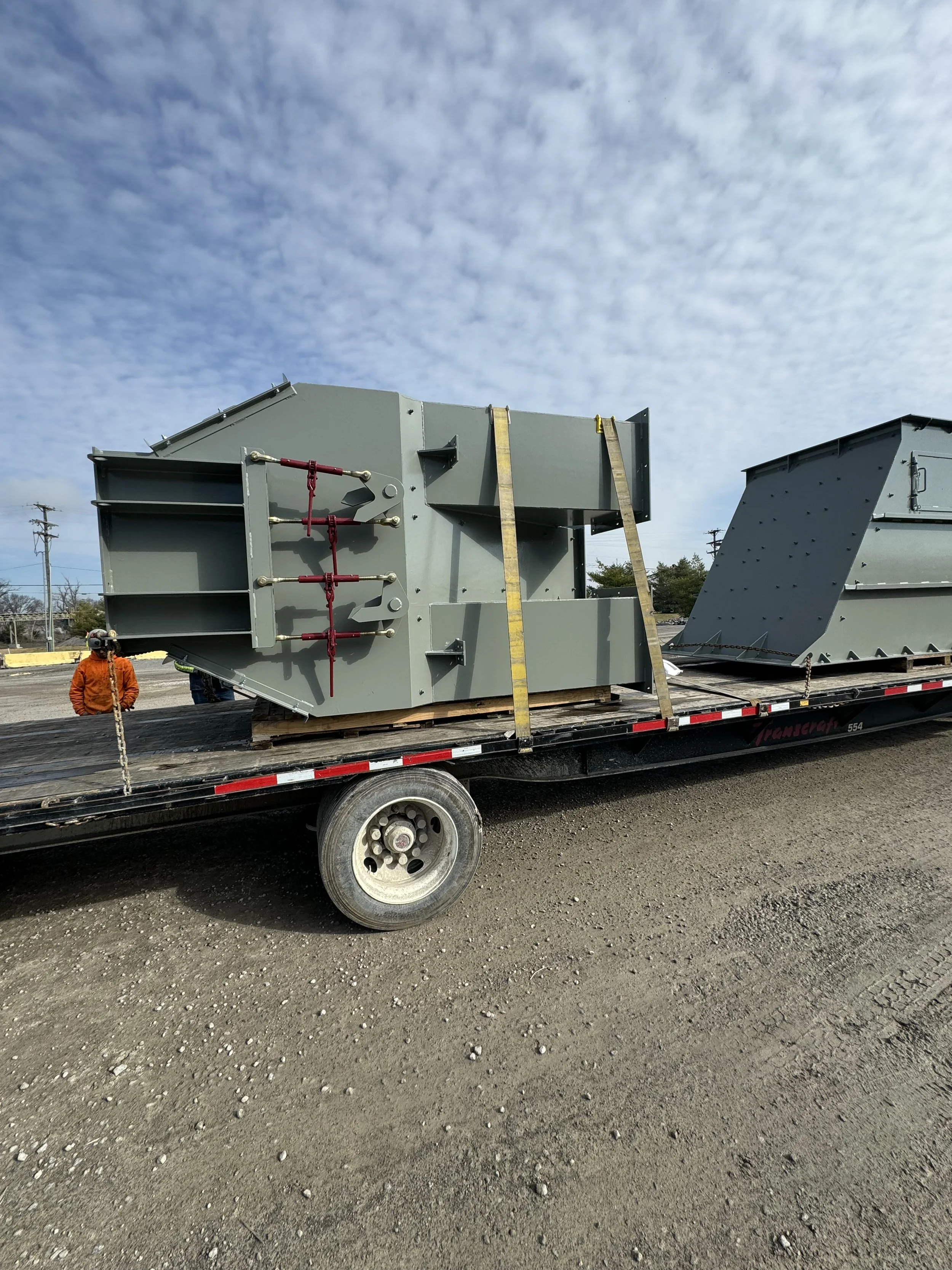 Flatbed truck carrying large gray industrial equipment in an outdoor setting, with workers standing nearby and a cloudy sky overhead.