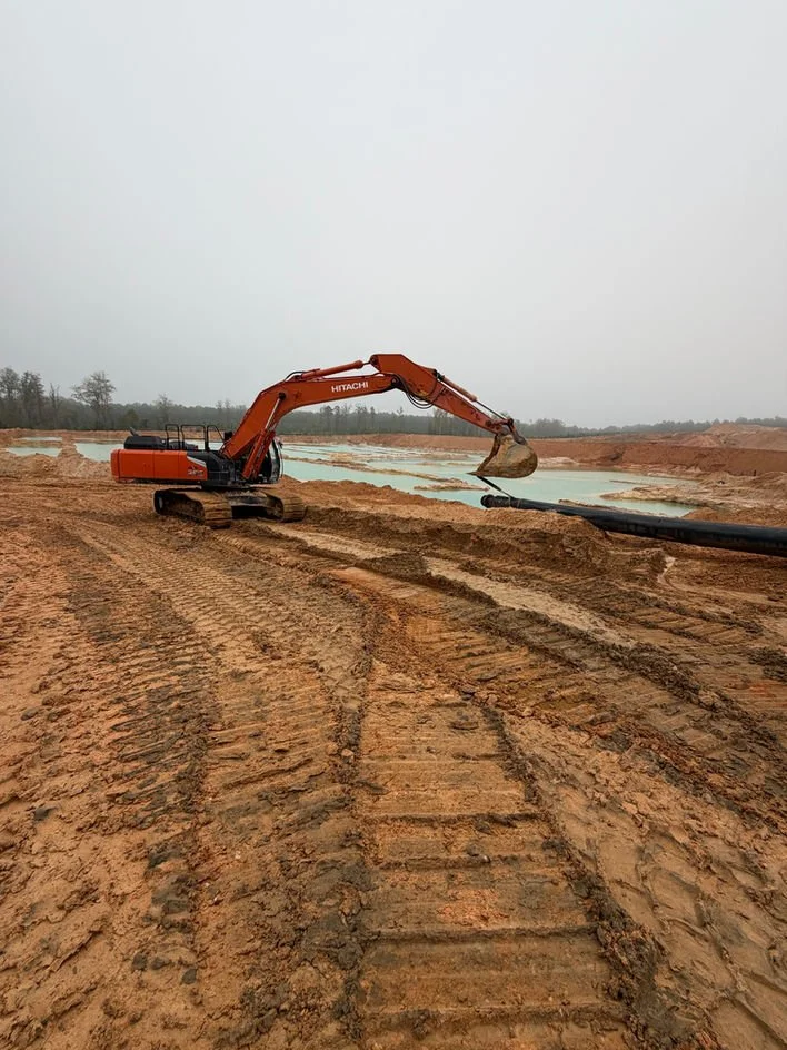 An orange excavator working on a construction site with large pipes, dirt tracks, and a water body in the background.