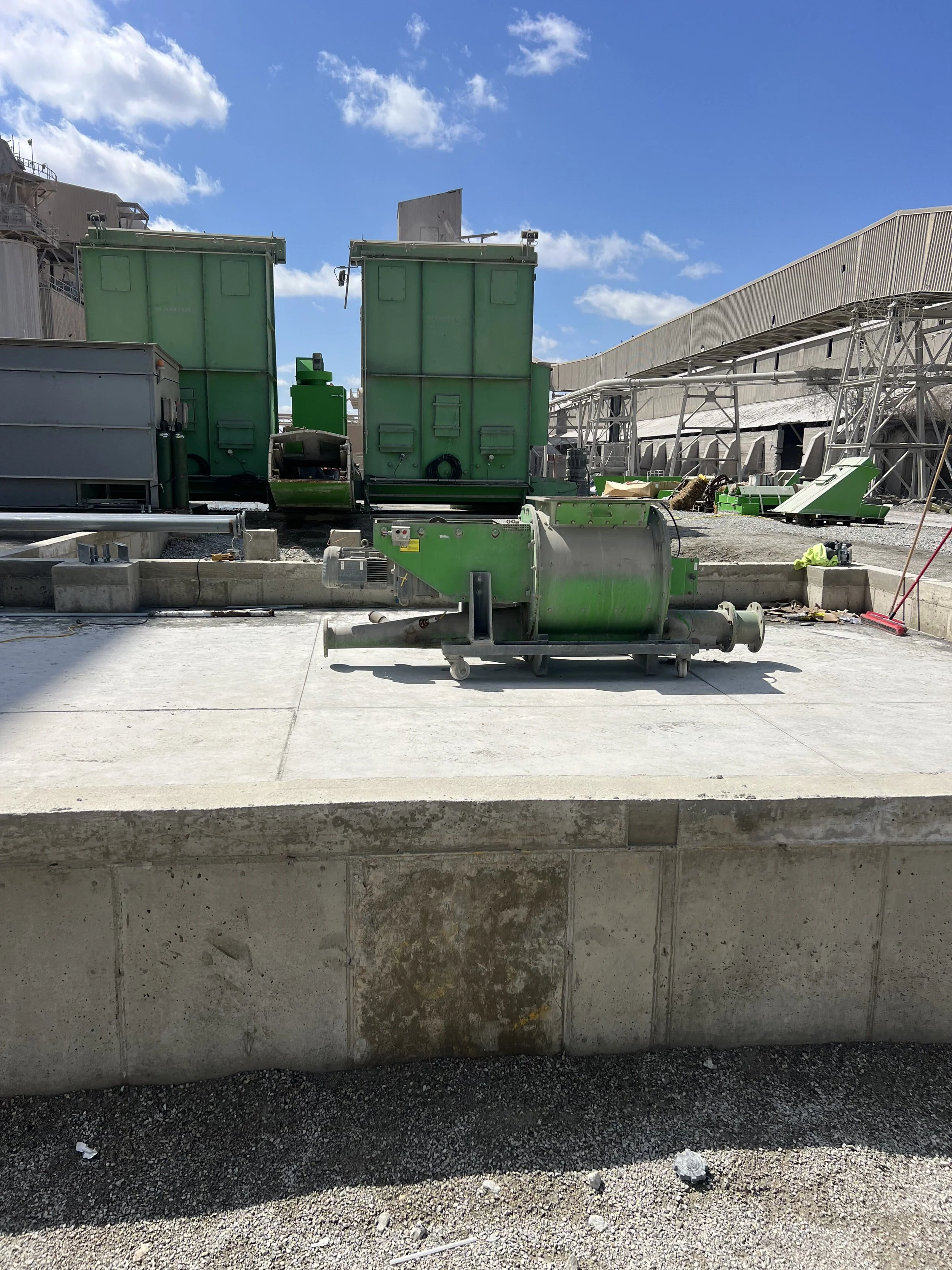 Construction site with green and gray industrial equipment, a concrete wall in the foreground, and a building with metal siding under a partly cloudy blue sky.