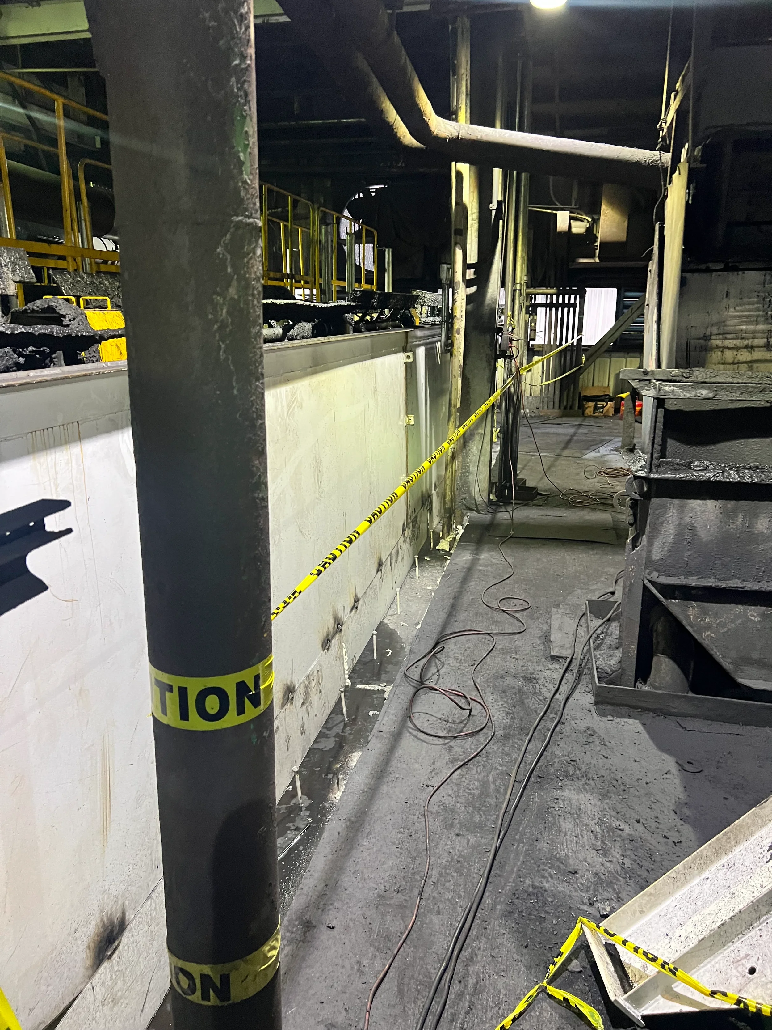 A scene at an industrial construction site showing damaged pipes, yellow caution tape, and electrical cords on the floor, with safety markings on a post in the foreground.