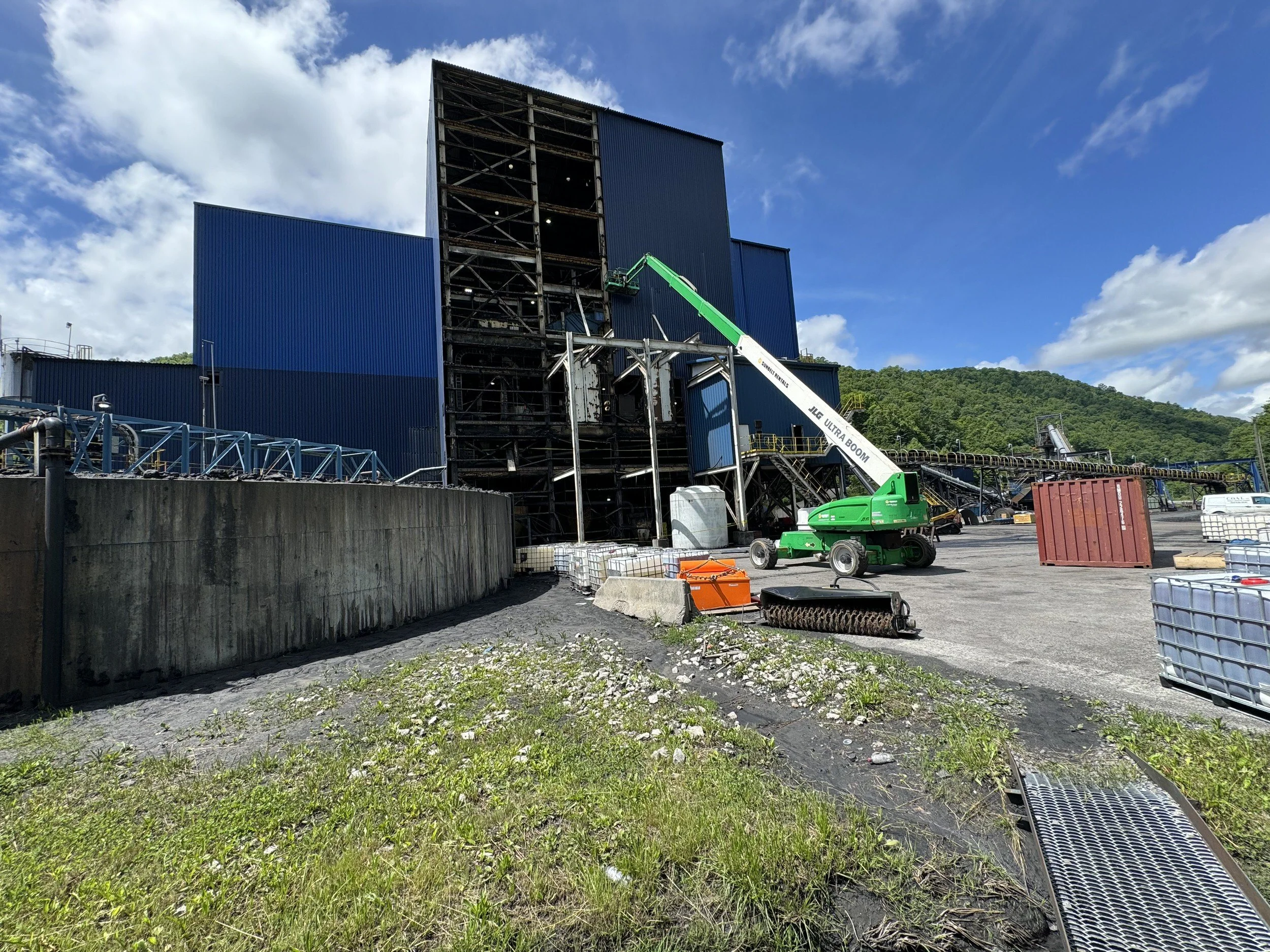 Industrial building under construction with blue exterior walls, a green cherry picker lift, and construction materials around, set against a background of hillside and blue sky with clouds.