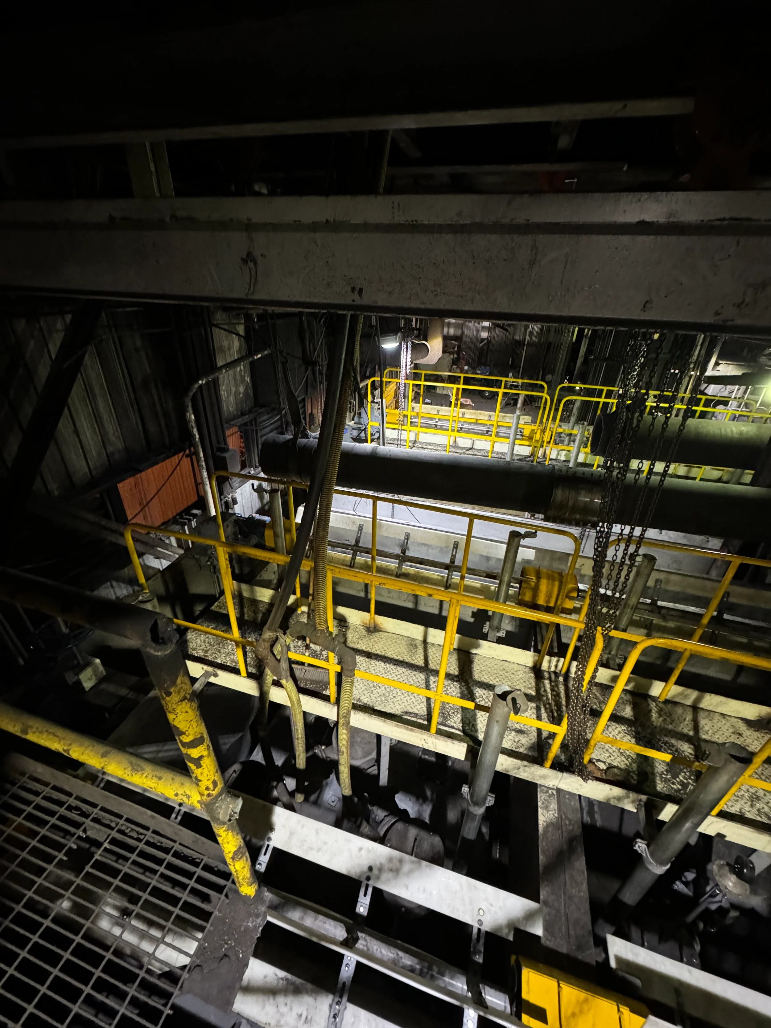 Industrial scene with yellow safety railings, pipes, chains, and metal grates in a dark, enclosed space.