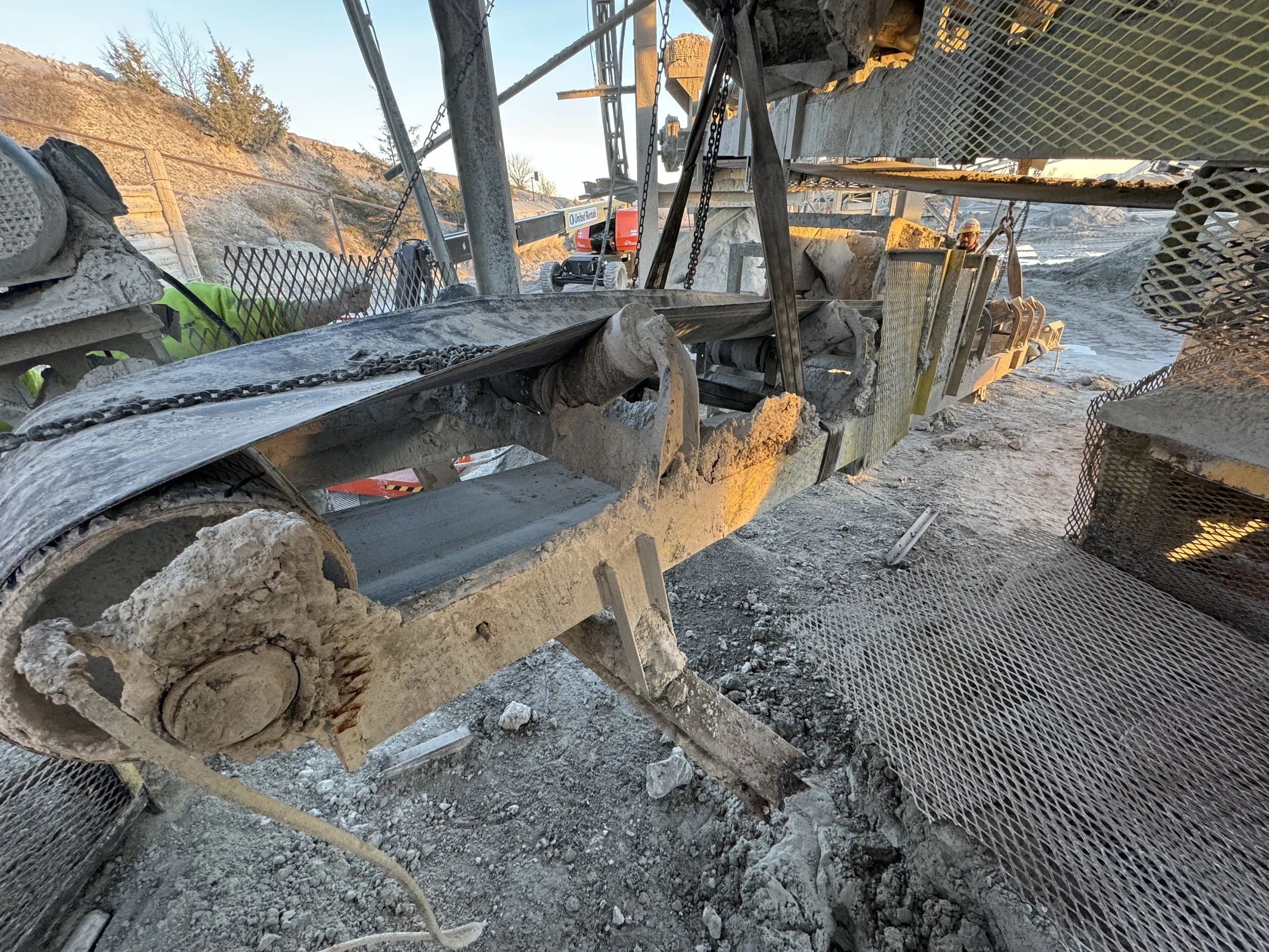 Construction worker in a hard hat operating heavy machinery at a rocky construction site. The machinery appears to be breaking or moving large rocks and debris. The environment is outdoors with hills and fencing in the background.
