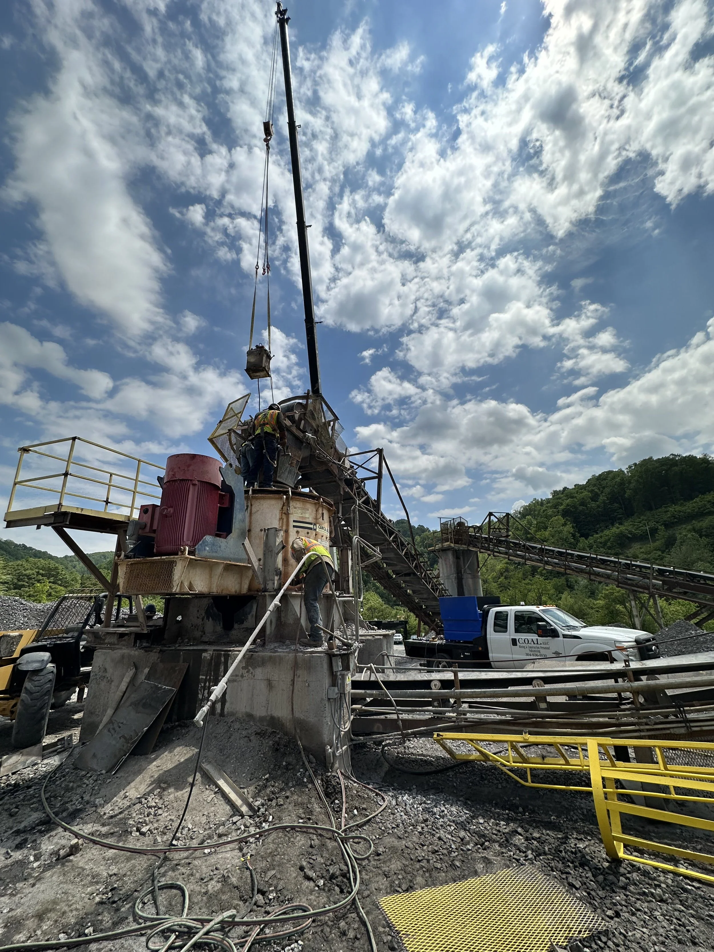 Workers operating heavy machinery at a construction site outdoors during daytime, with a cloudy sky and green hills in the background.