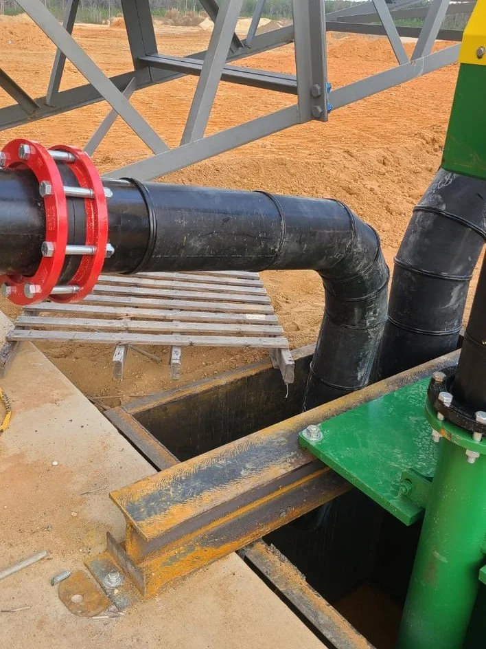 Close-up of industrial piping with a large black pipe connected to a valve and a green support, set on a construction site with orange dirt and metal scaffolding in the background.
