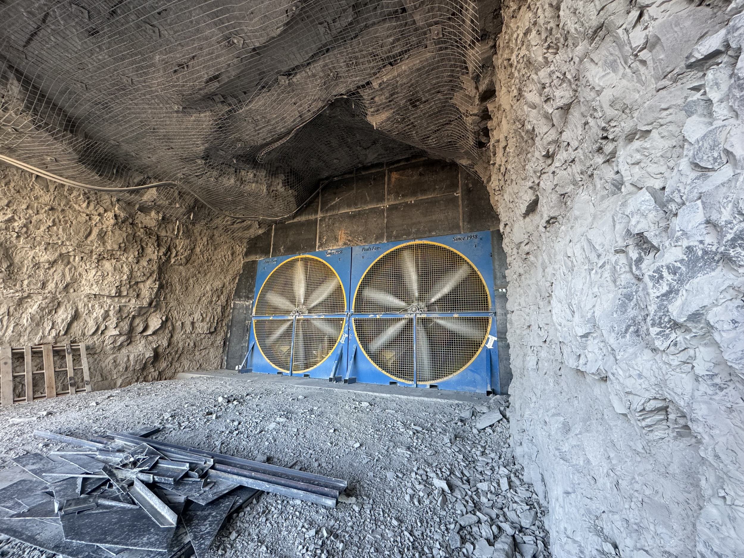 Construction inside a rocky tunnel with a large industrial fan and construction materials on the ground.