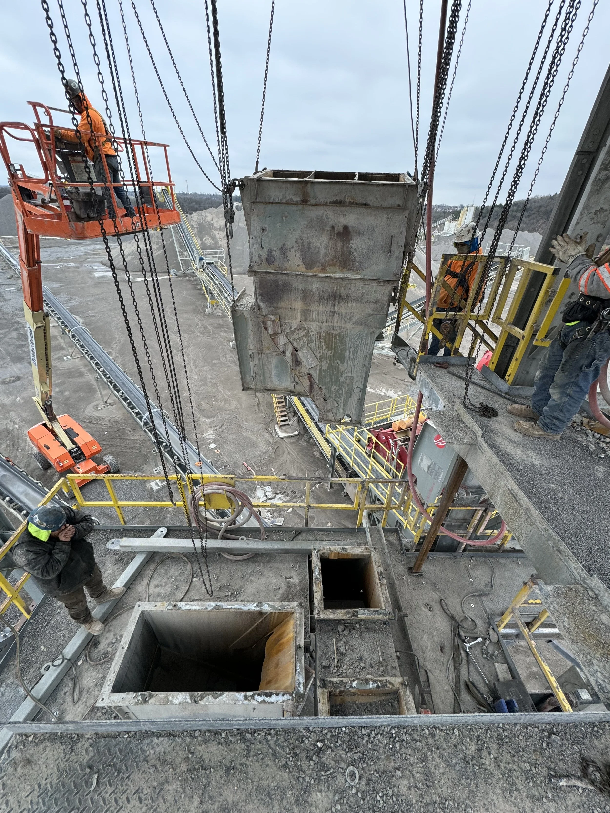 Construction workers working on a site, lifting large metal structures with cranes, with construction equipment and trenches visible.