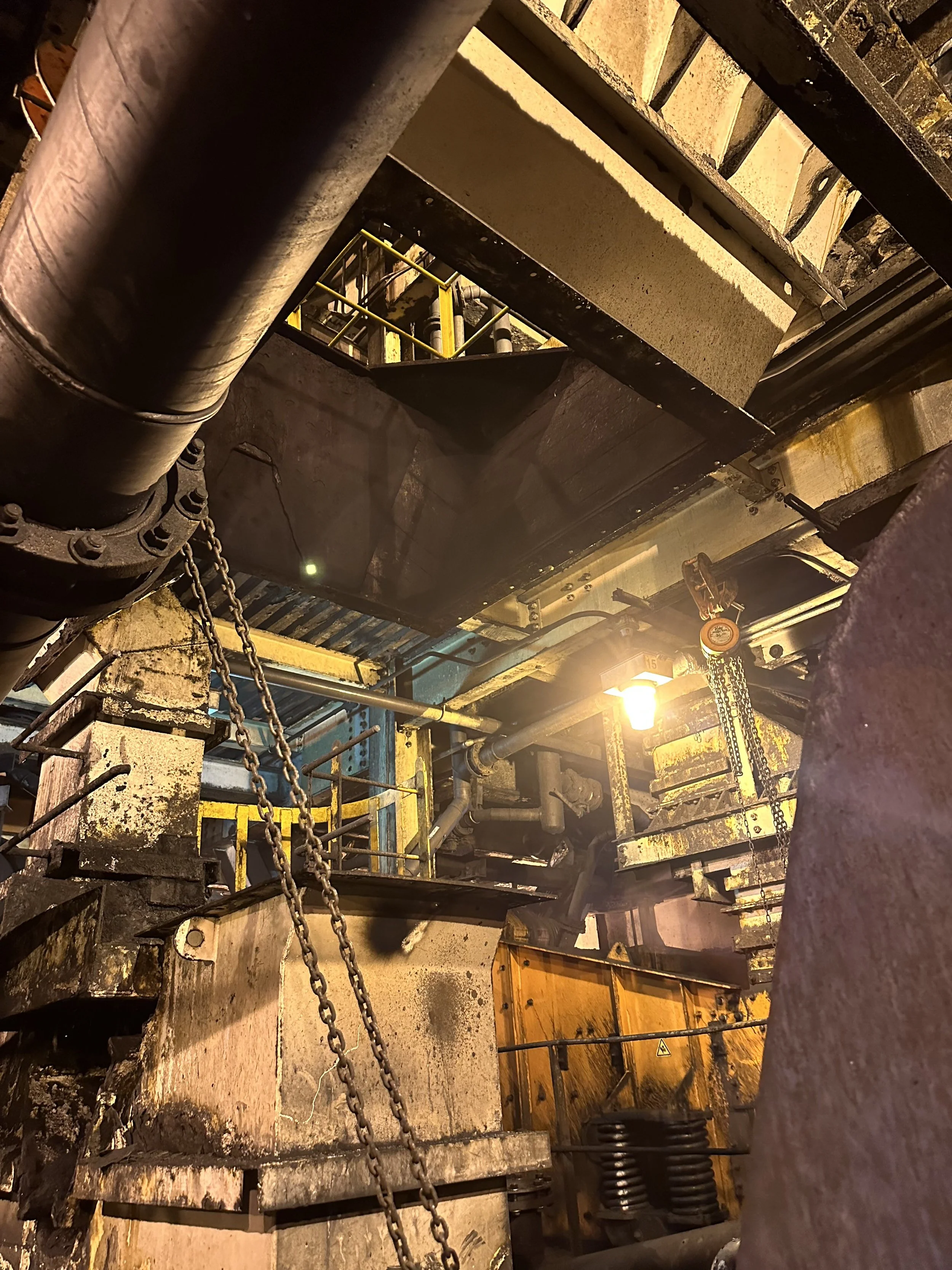 Industrial machinery and metal components, including chains, pipes, and yellow safety barriers, in a factory or workshop setting with overhead lighting.