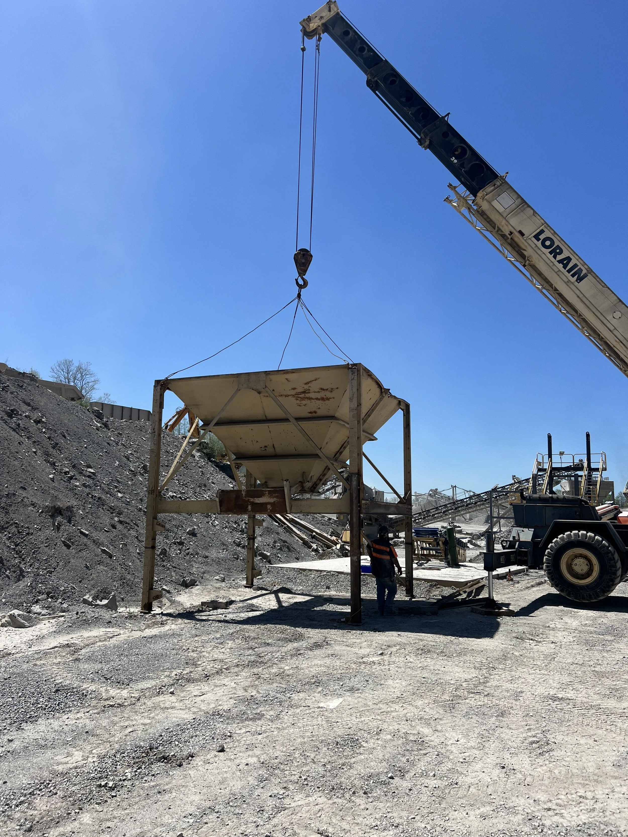 A construction site with a crane lifting a large, metal container under a clear blue sky. There is a worker standing nearby, and various construction equipment is visible in the background.