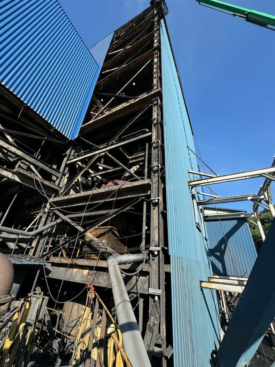 A tall industrial building with blue metal siding, exposing rusted metal framework and pipes inside, under a clear blue sky.