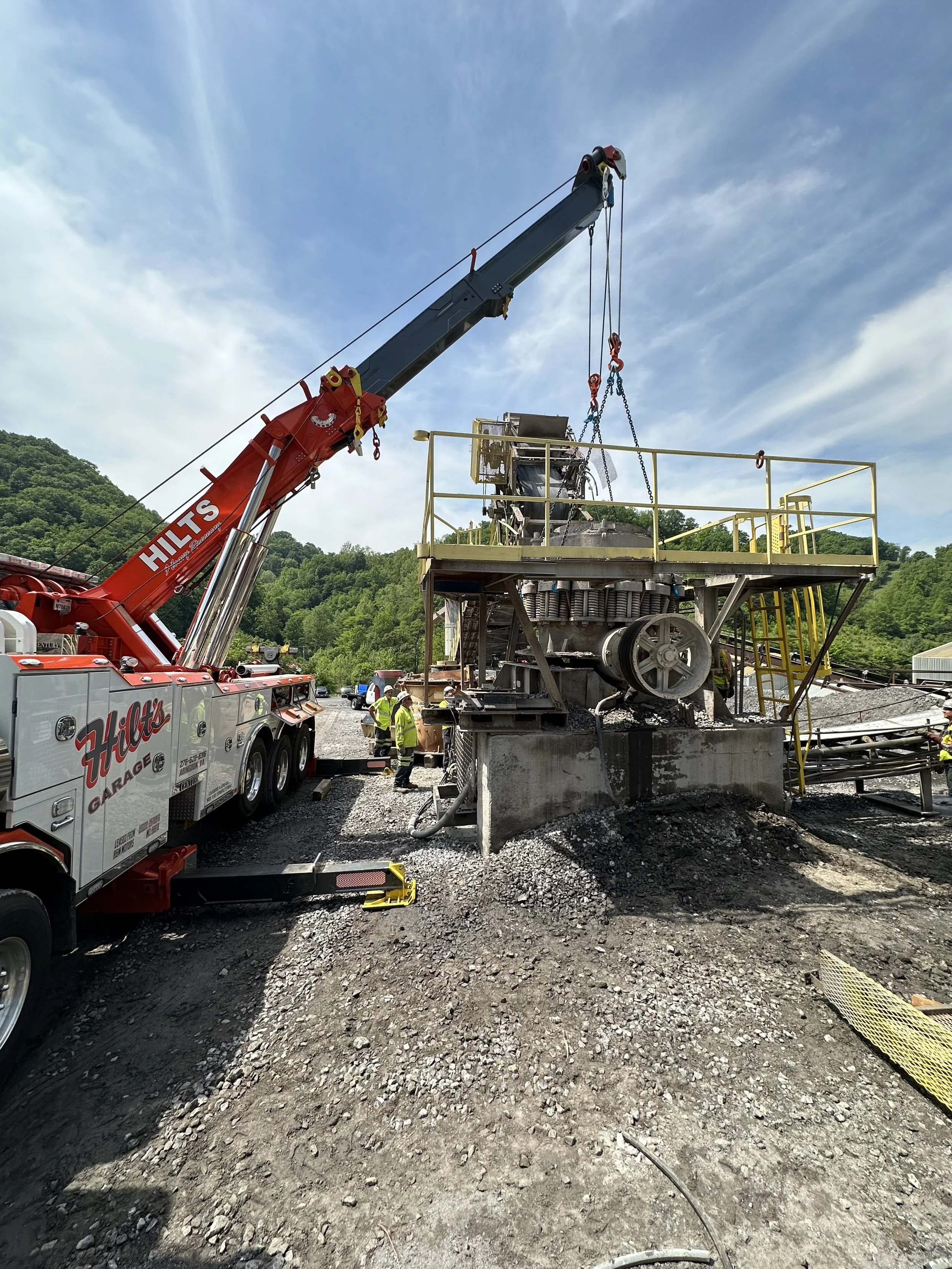 Construction site with a large crane lifting parts of a machine, multiple workers in safety gear, and a mountainous background under a partly cloudy sky.