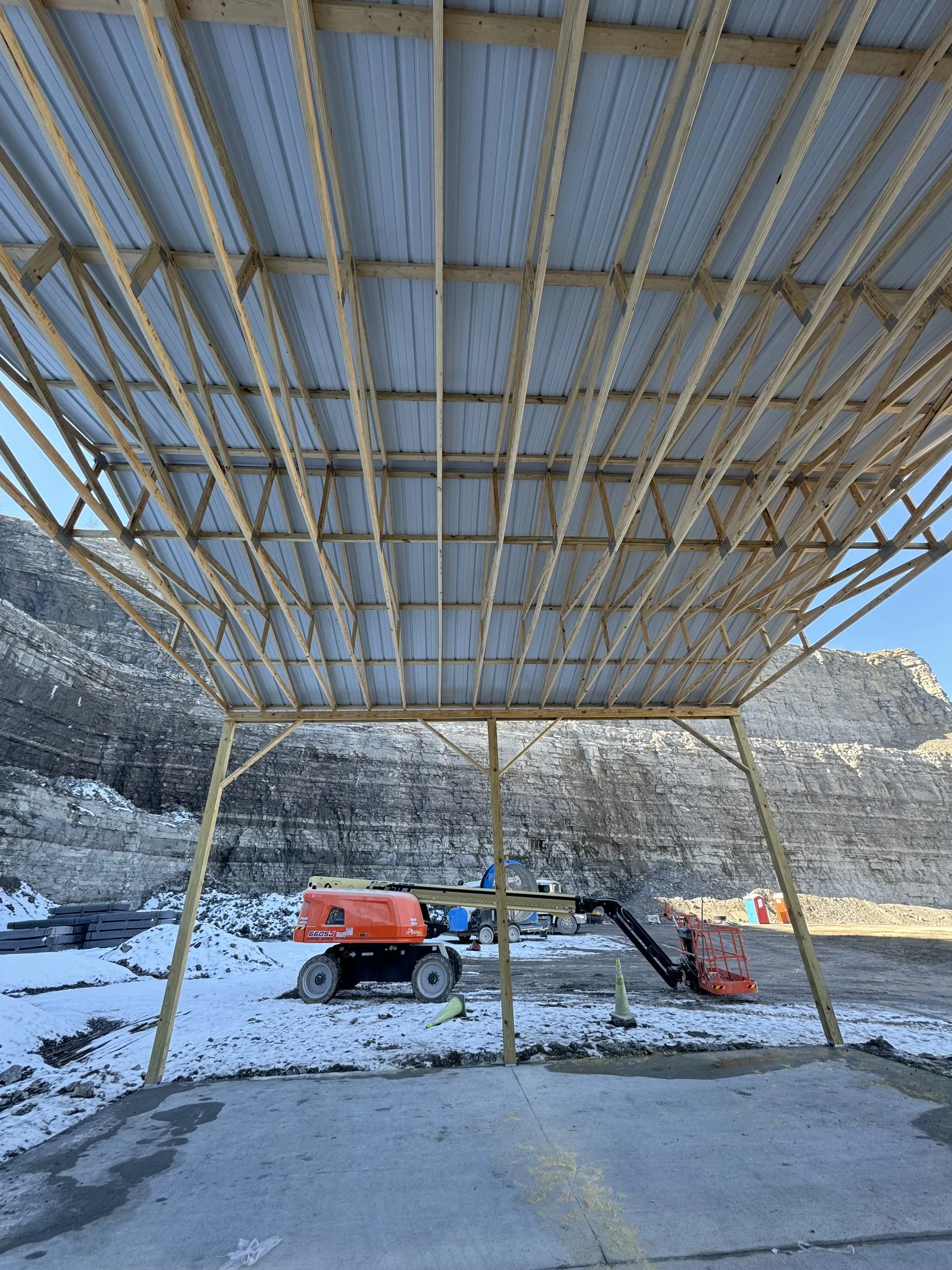 Construction site featuring a wooden framework for a new structure, with a metal roof installed. Construction equipment and orange safety cones are visible on the snowy ground, with rocky cliffs in the background.