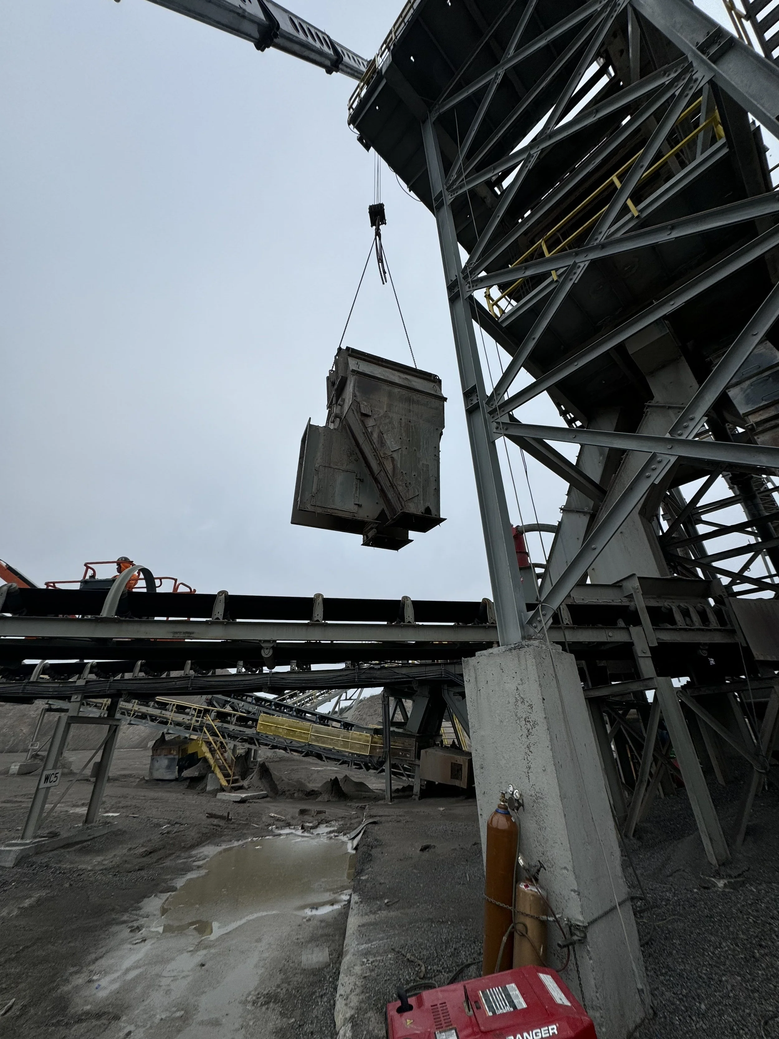 Industrial construction site with metal framework, conveyor system, and construction worker in orange safety vest, with cloudy sky in background.