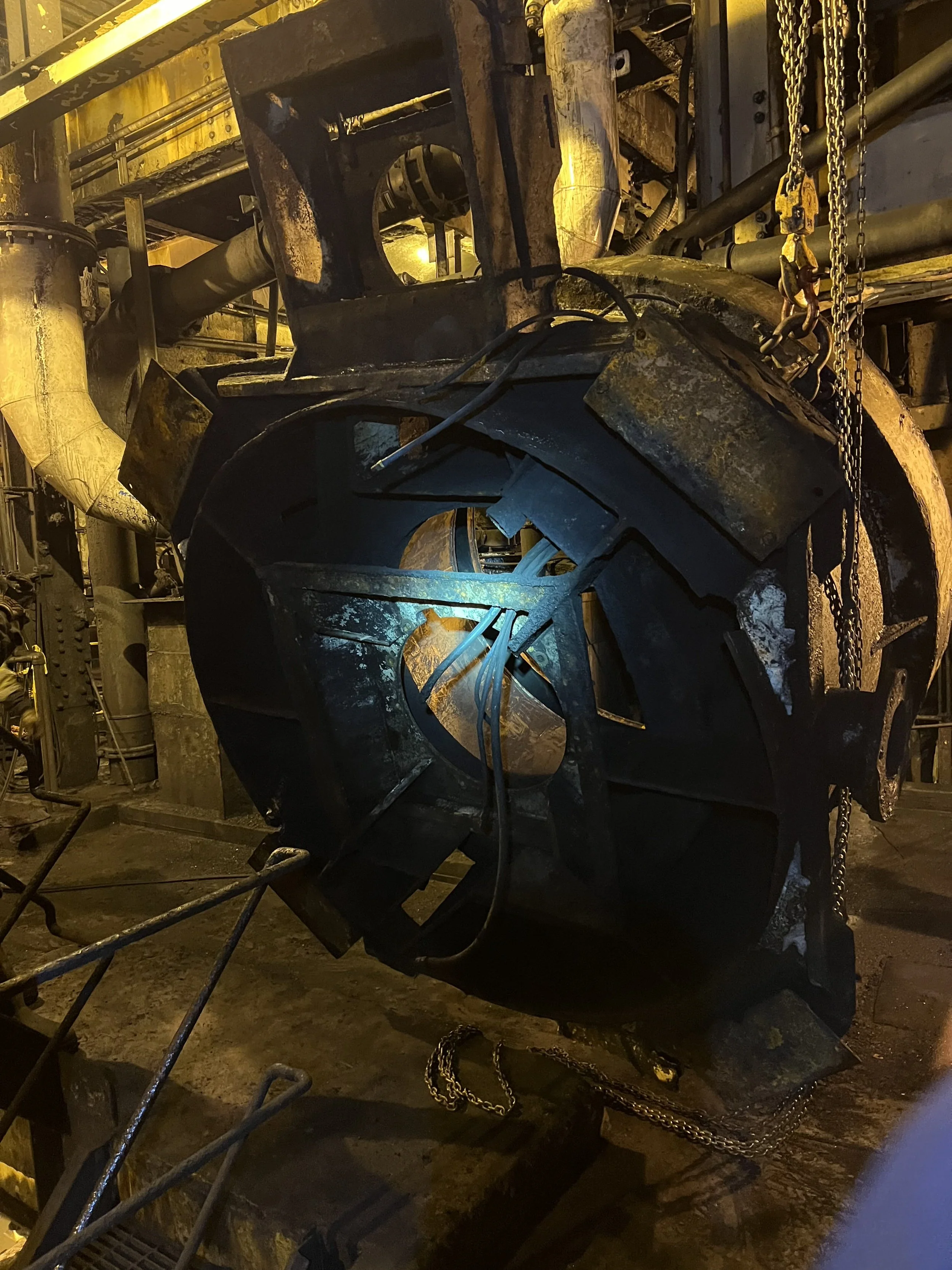 Close-up view of an industrial or factory setting with machinery, pipes, chains, and metal components, some of which are rusted and damaged, with yellow lighting in the background.