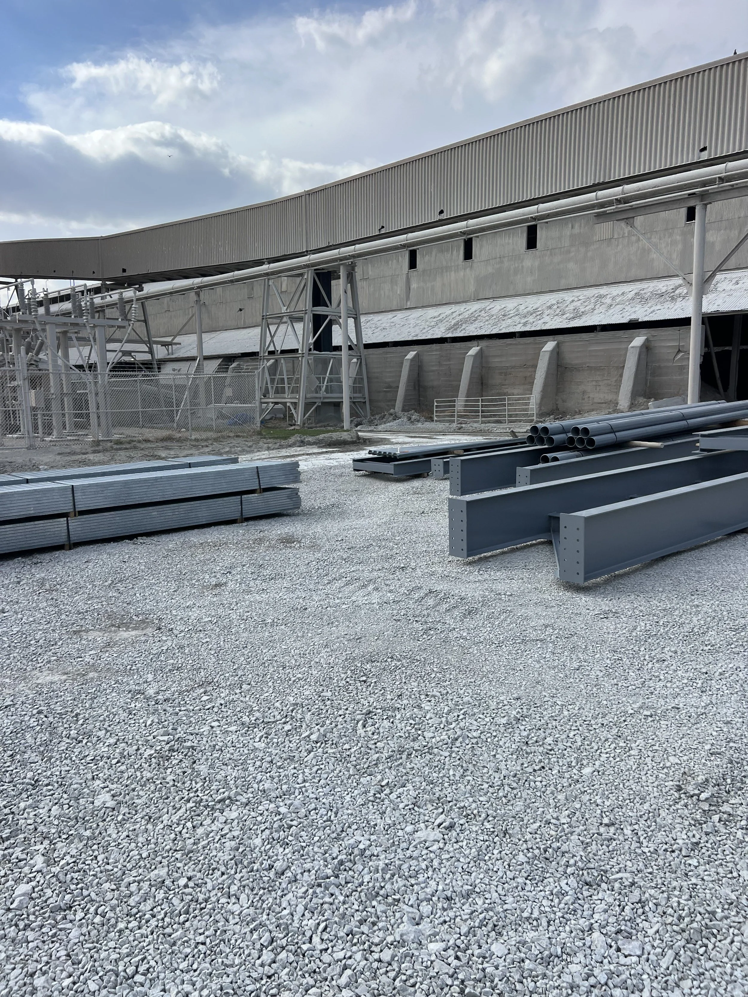 Construction site with metal pipes, structural supports, and gravel ground under a partly cloudy sky.