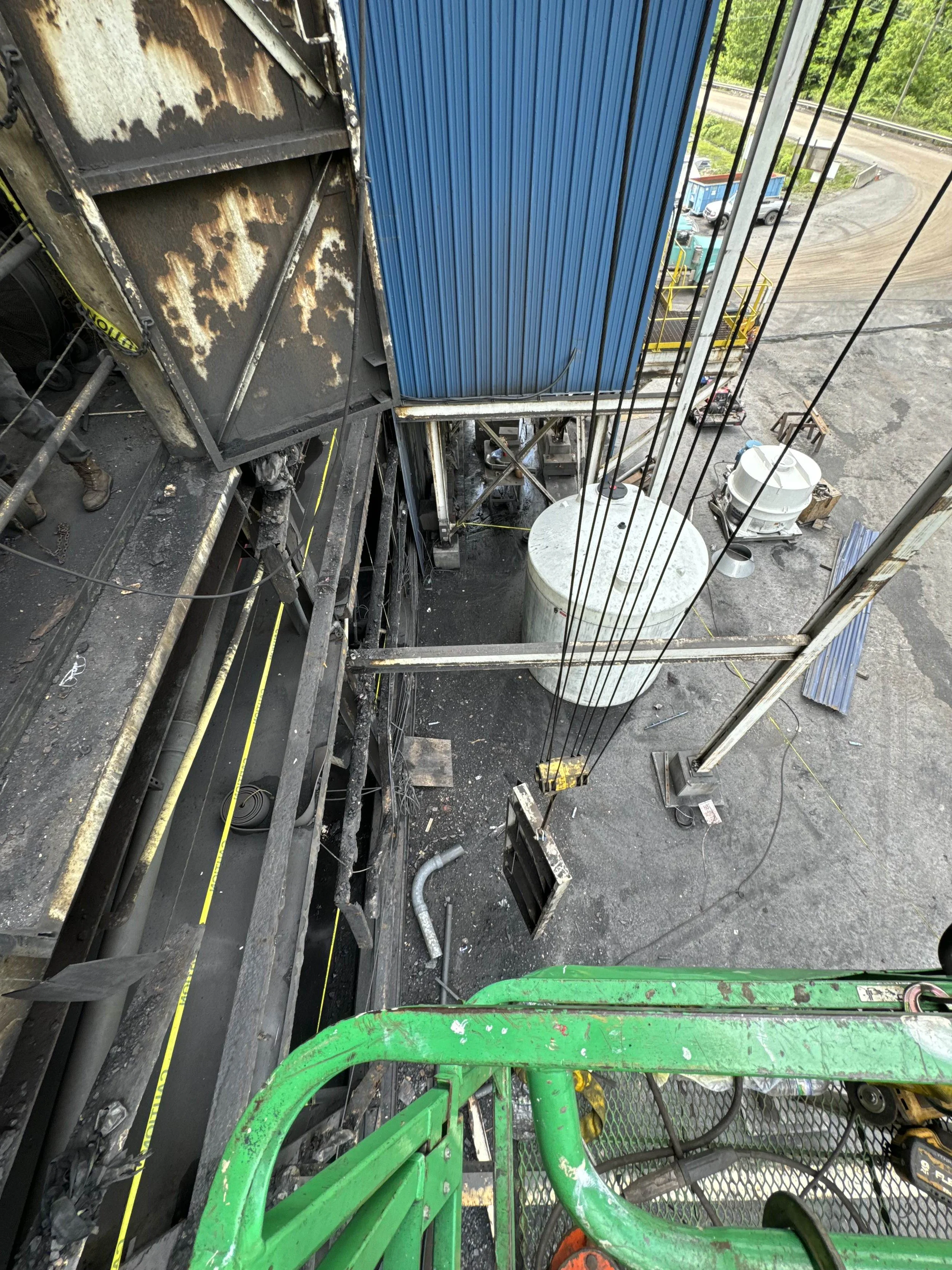 View from a lift showing construction work on a building with rusted metal and a blue panel. The ground below is a construction site with dirt, pipes, and equipment. Green and orange safety equipment are visible.
