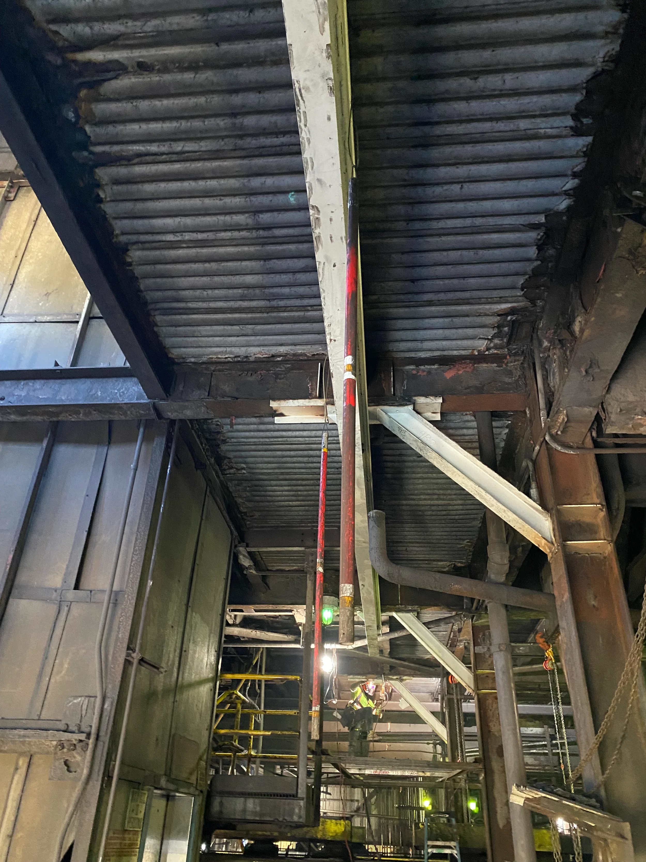 Construction workers on scaffolding working on a ceiling with metal insulation panels, pipes, and electrical wiring in an industrial building.
