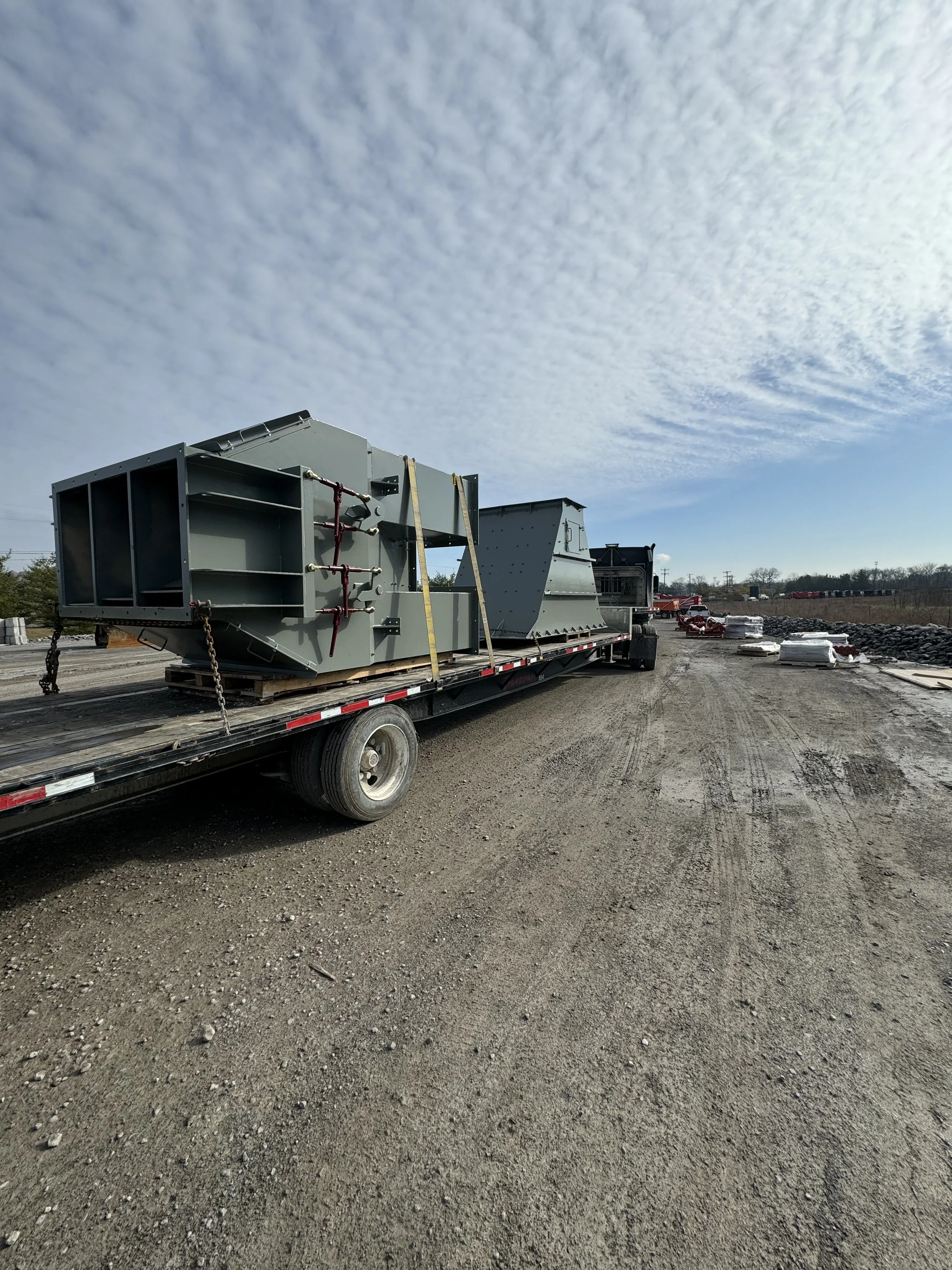 A flatbed truck carrying large metal industrial equipment in an outdoor lot. The sky is partly cloudy and there are additional construction materials and equipment in the background.