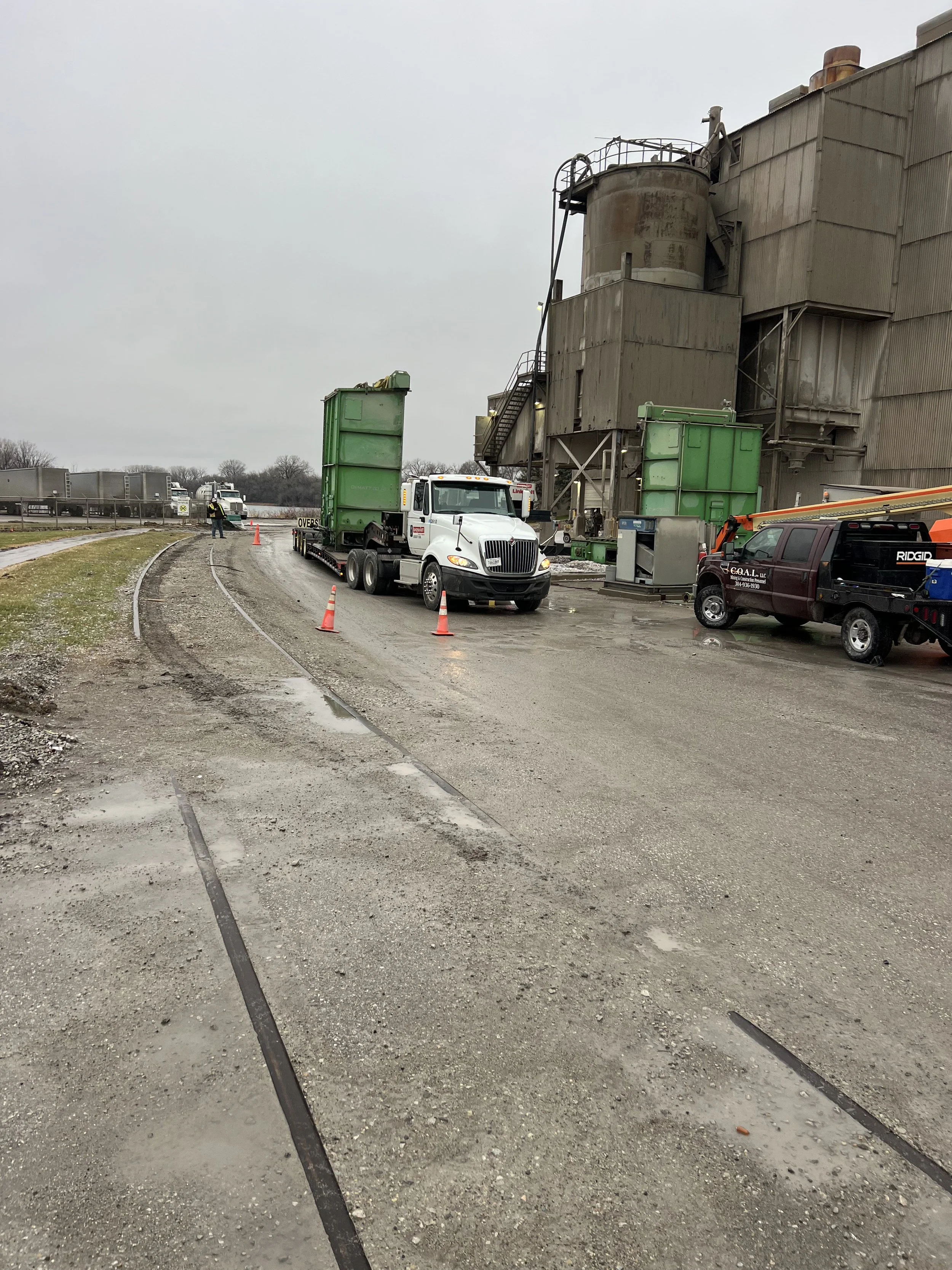 Road construction or maintenance scene with trucks, traffic cones, and workers near an industrial building on a cloudy day.