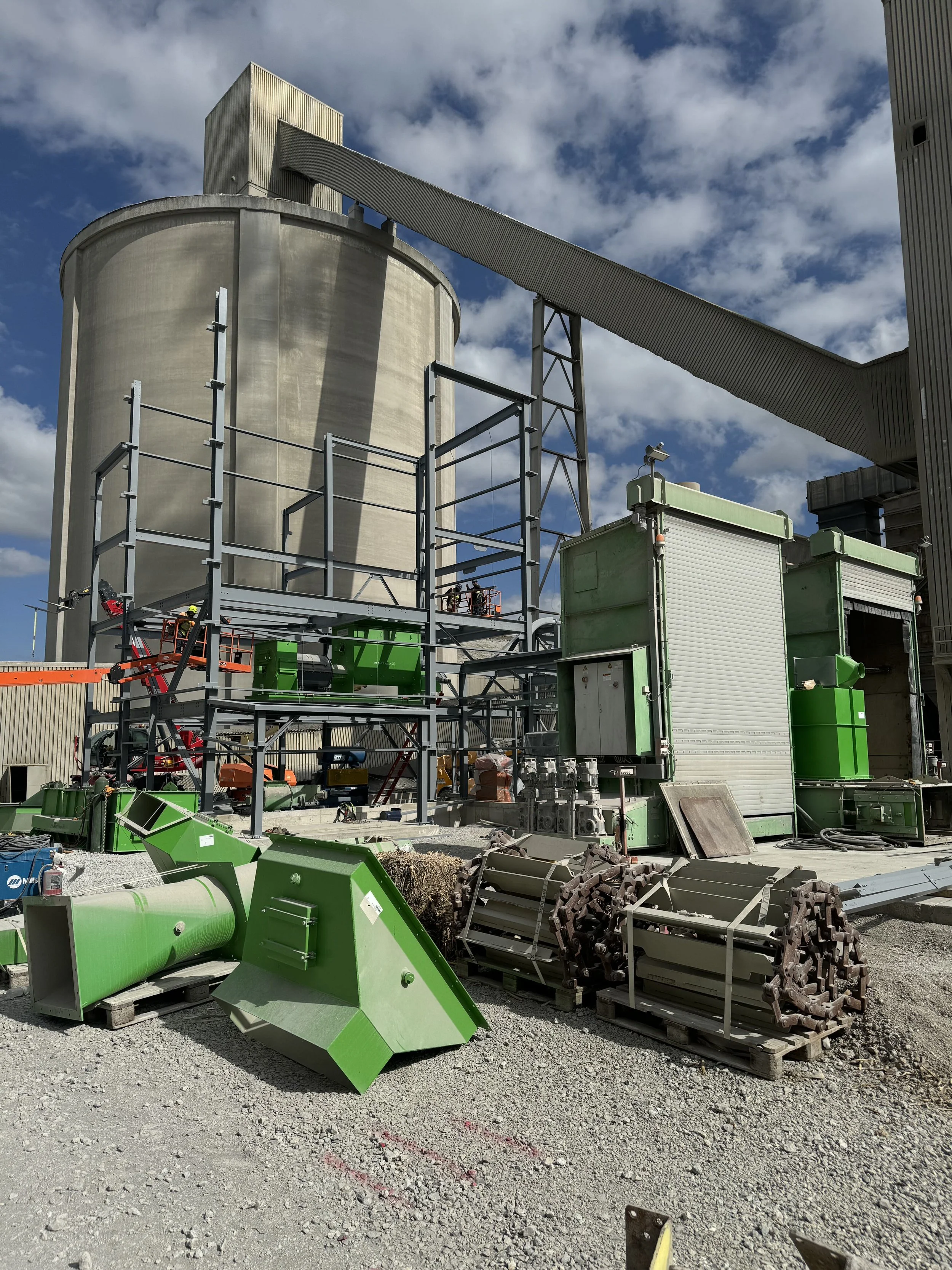 Construction site with large concrete silo, green construction equipment, and various metal and machinery parts scattered on gravel ground.