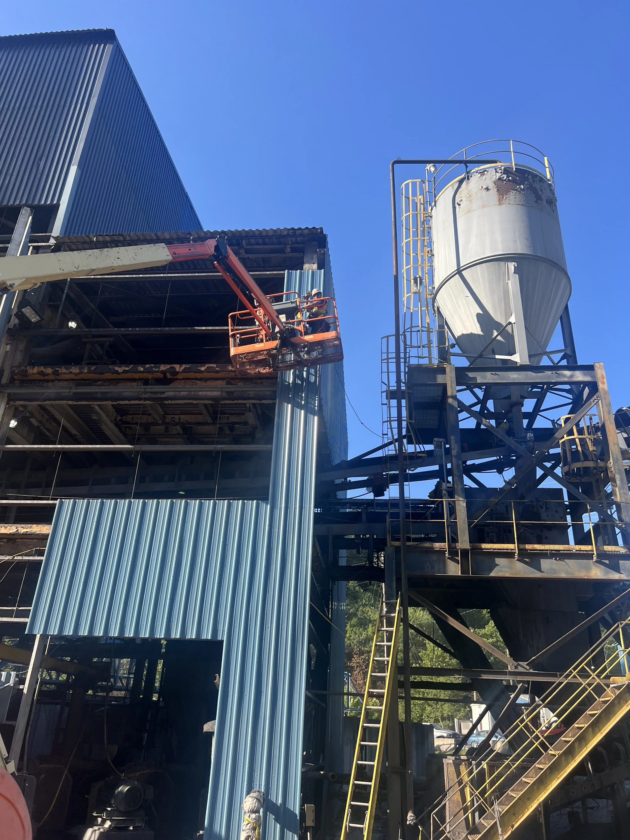 Steel building under construction with workers on a scissor lift, a large silo, and yellow safety railings against a clear blue sky.