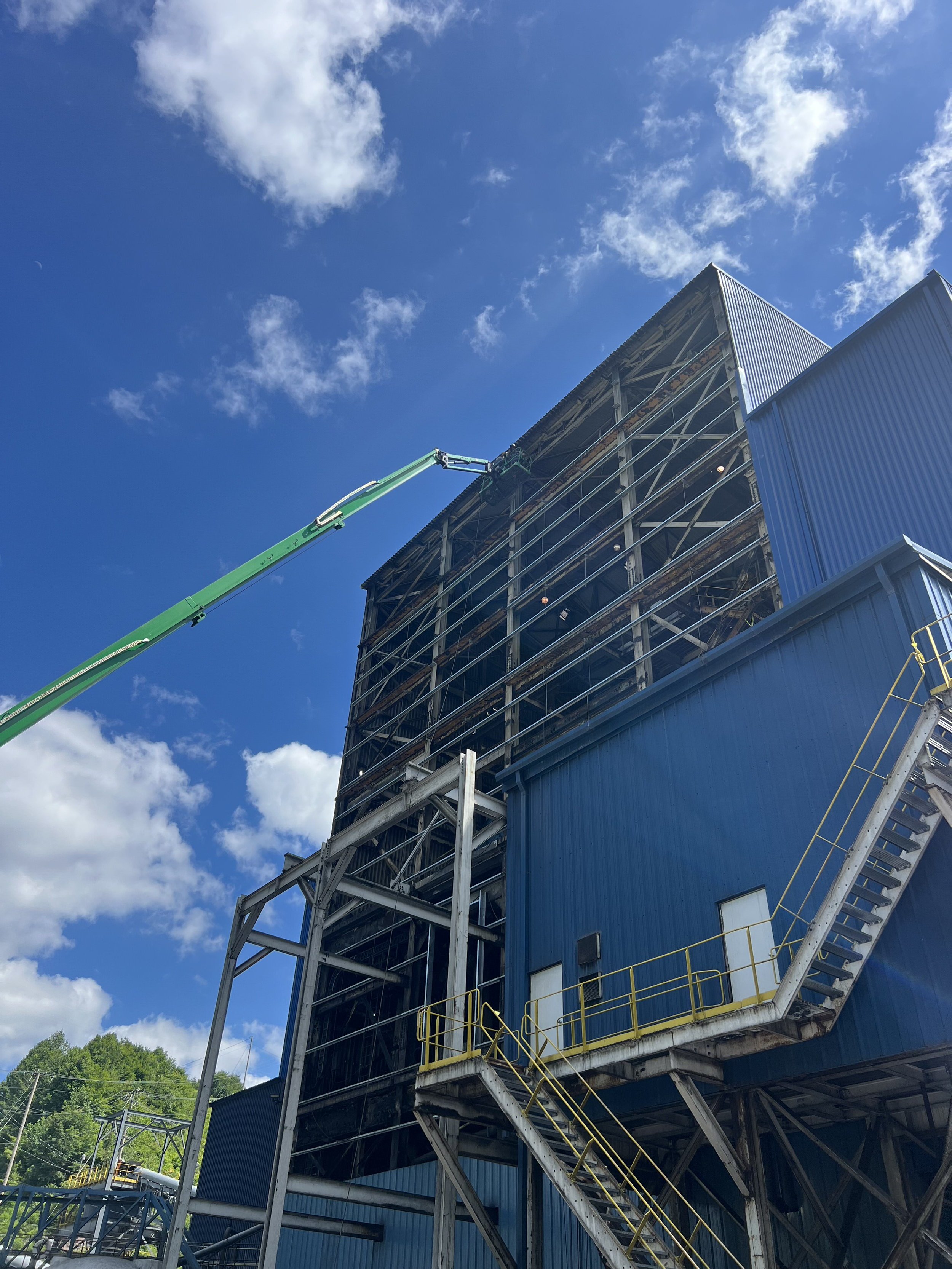 A large industrial building with blue and metal exterior, scaffolding, and a green crane arm extending toward the building, set against a blue sky with white clouds.