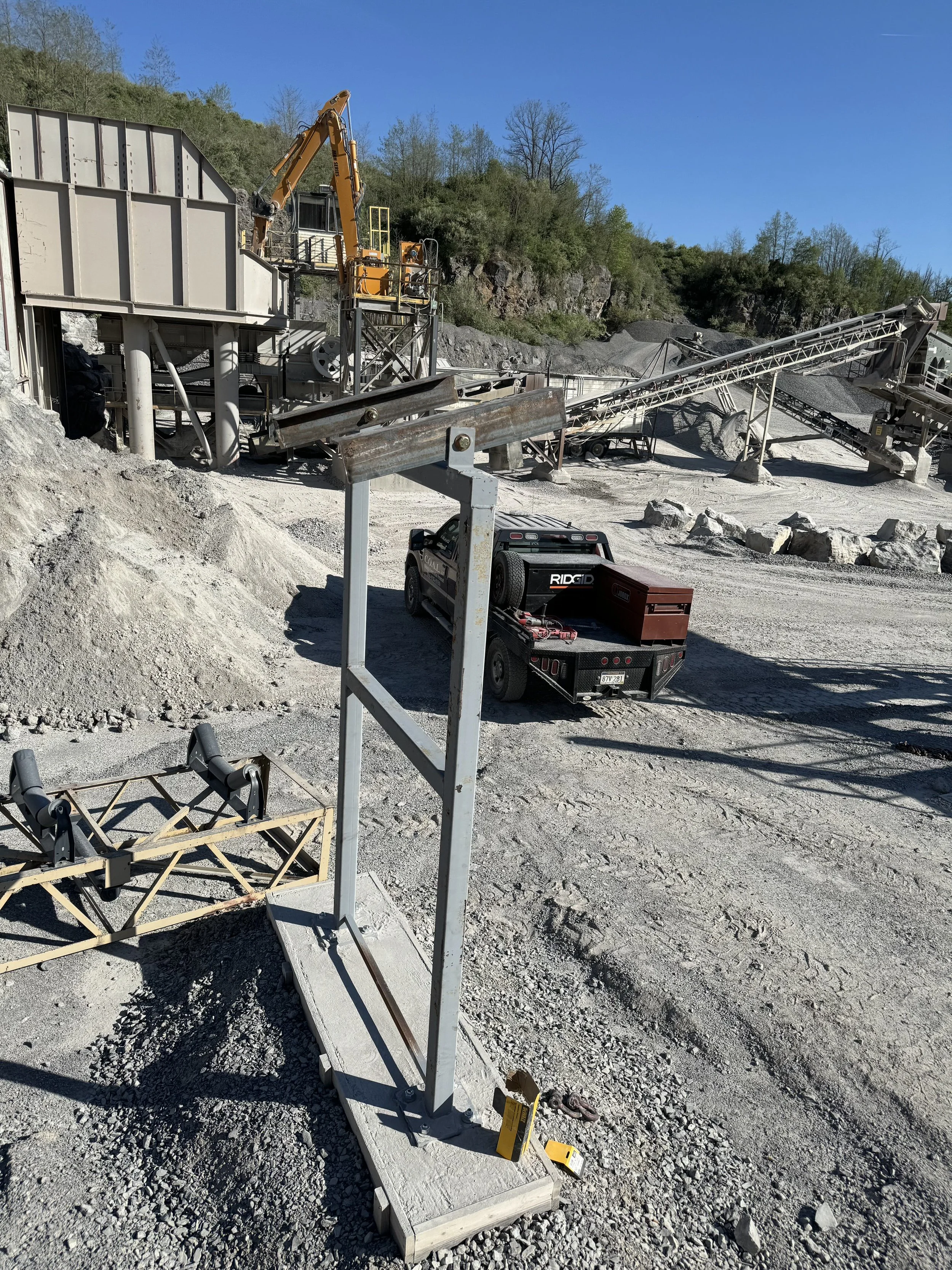 Construction site with machinery, gravel ground, and surrounding hills and trees, under a clear blue sky.