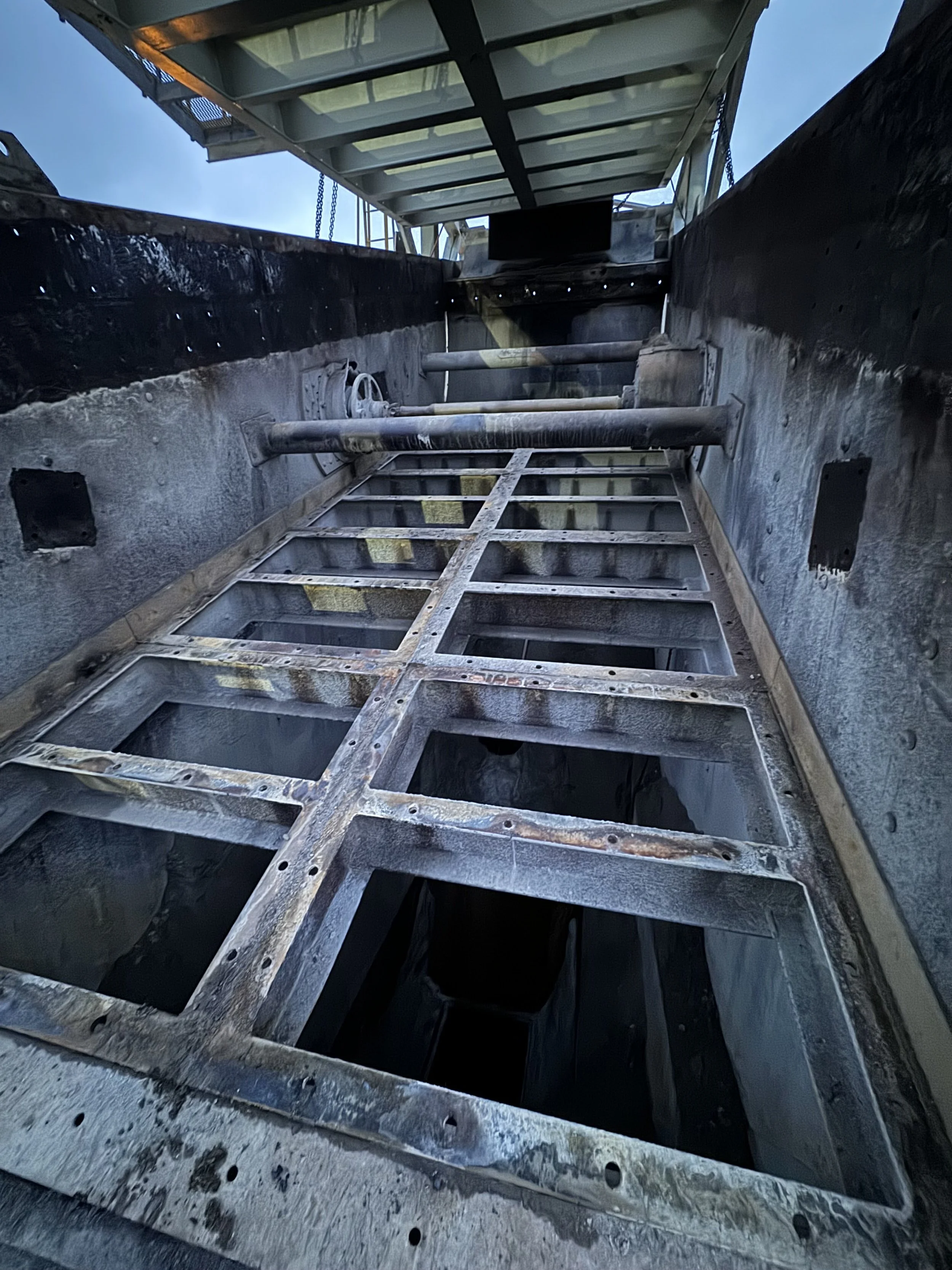 Looking up inside a metallic ship or aircraft structure with exposed beams, pipes, and a ladder, under a clear blue sky.