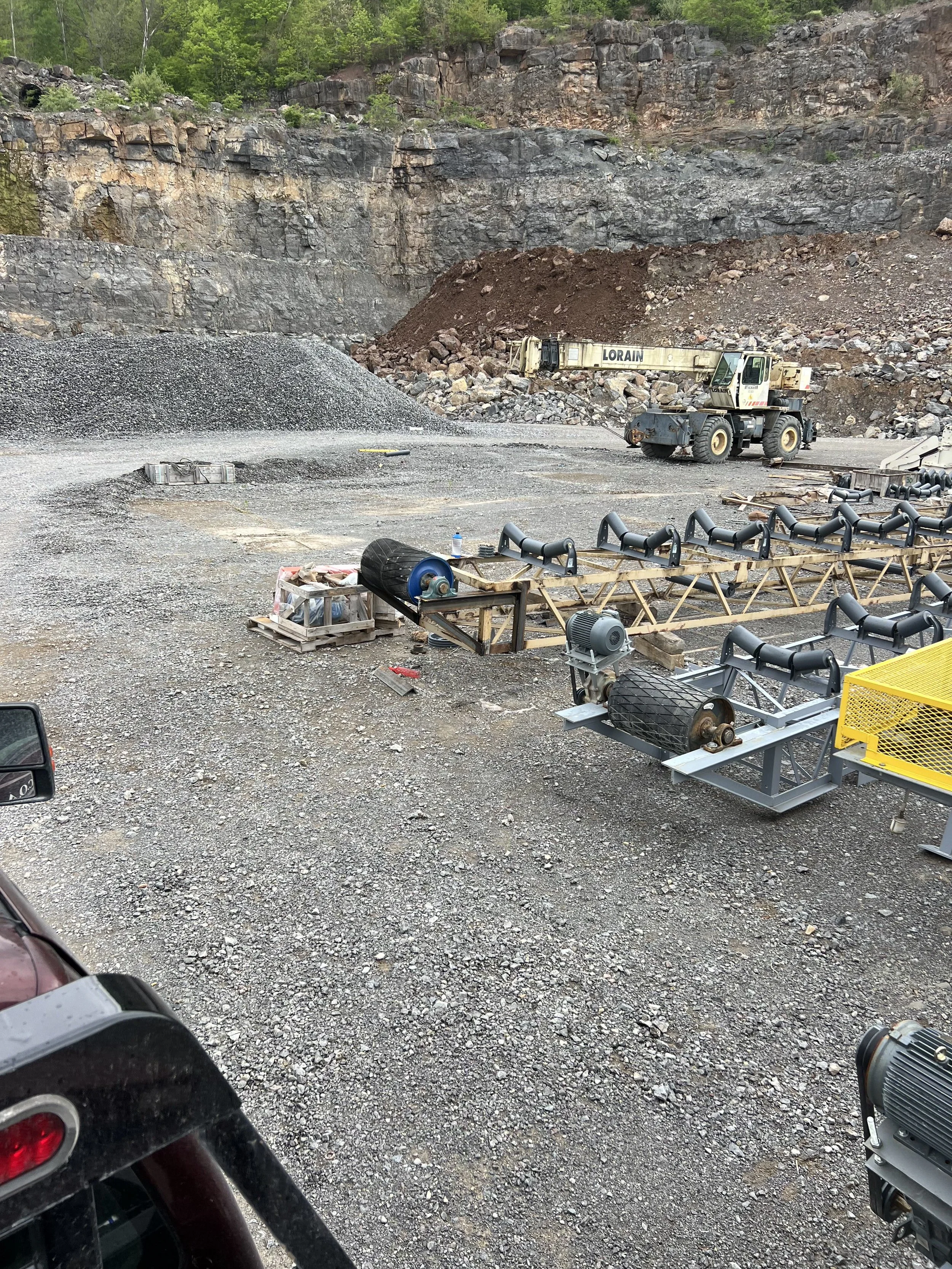 A construction or industrial site in a rocky outdoor area with a steep cliff face composed of layered rocks and green trees at the top. In the foreground, there are various pieces of equipment and machinery, including what appears to be a small conve