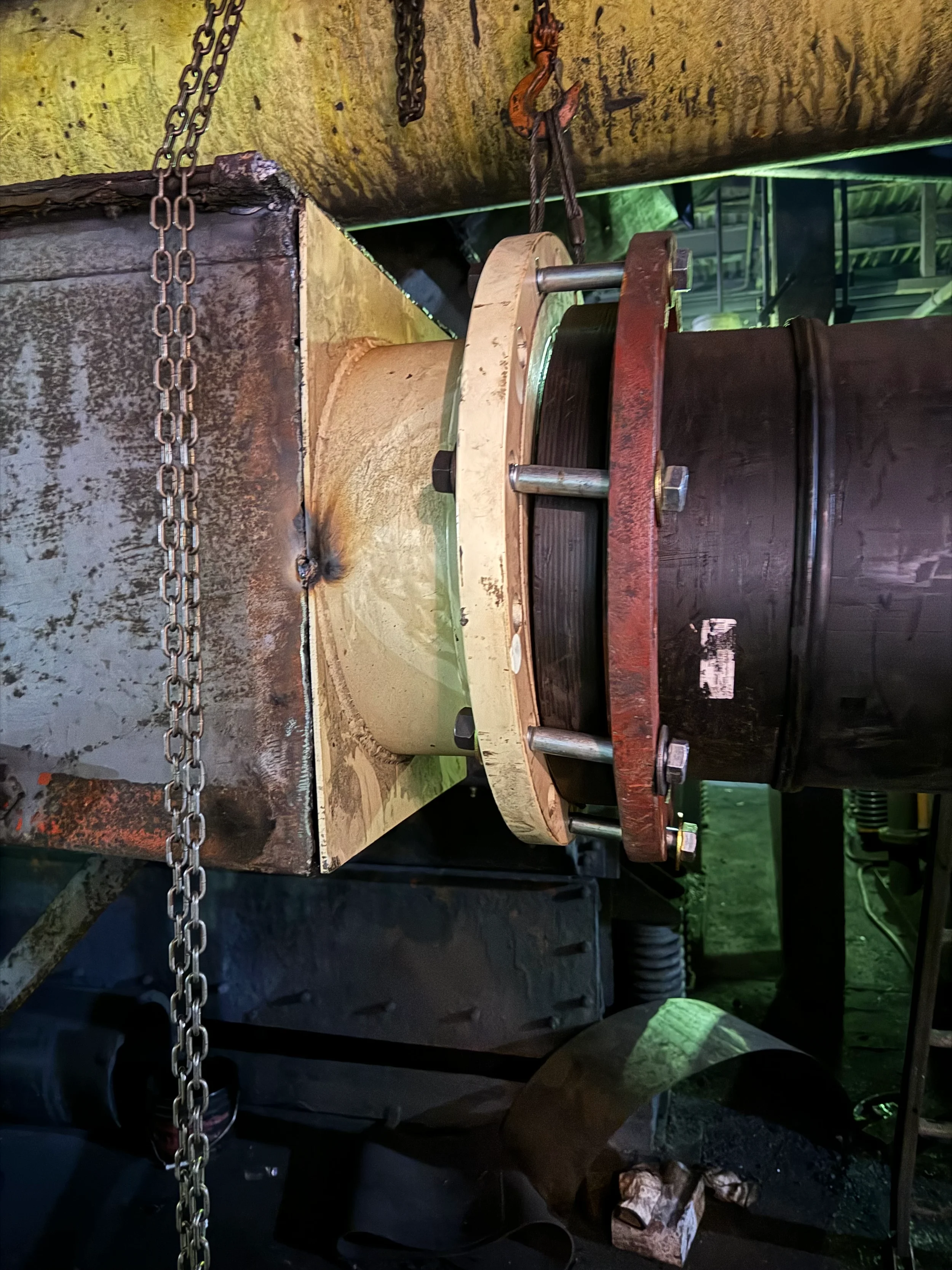 A close-up of a large industrial machine component with a chain hanging from the top left, a rusty hook, and bolts securing a large black and red drum or hopper. The machinery appears aged and stained with oil or grime, with a greenish light in the b