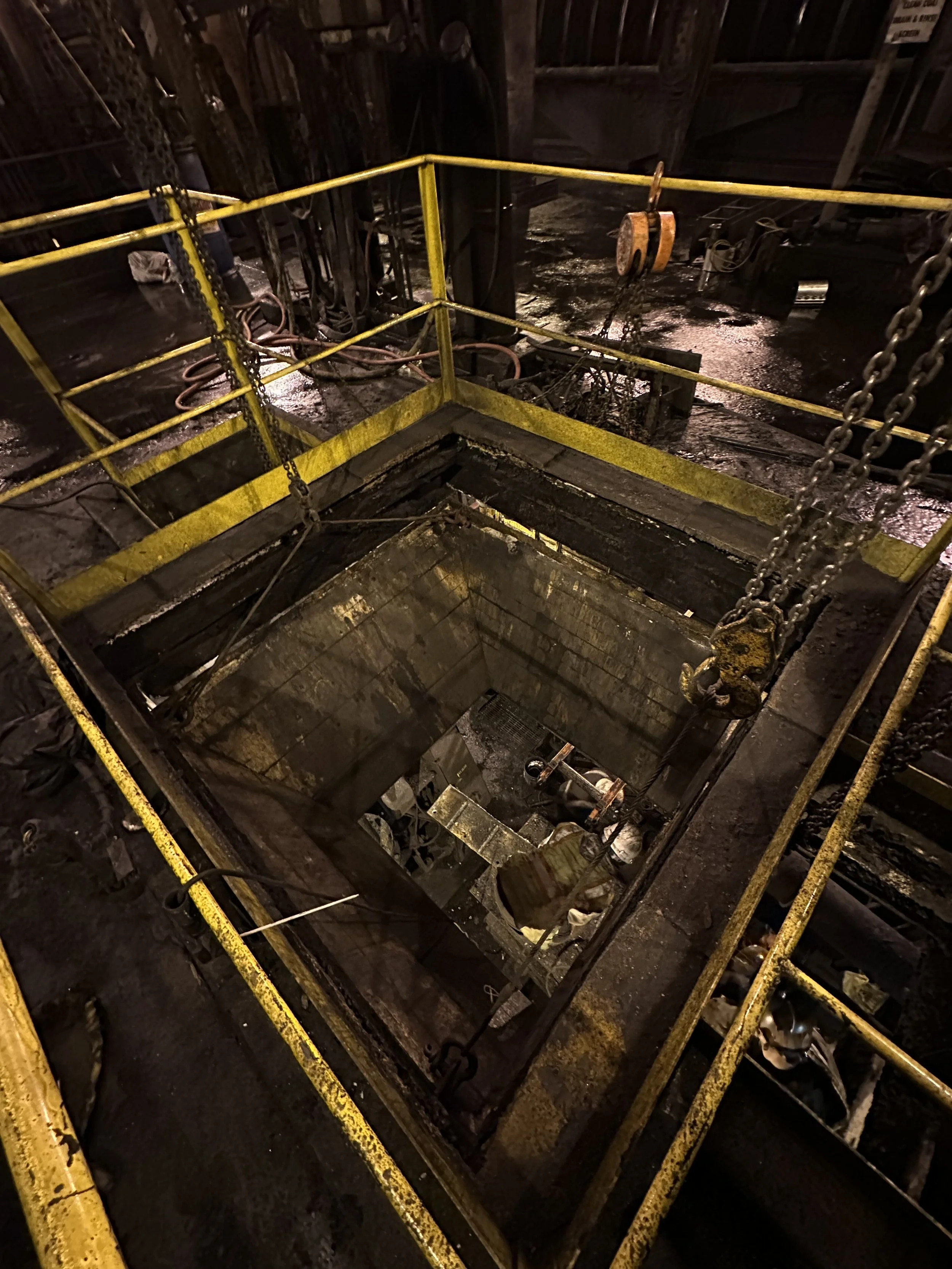 An industrial or construction site with a deep pit or excavation surrounded by yellow safety railings, chains, hooks, and equipment, with a dark and muddy environment.