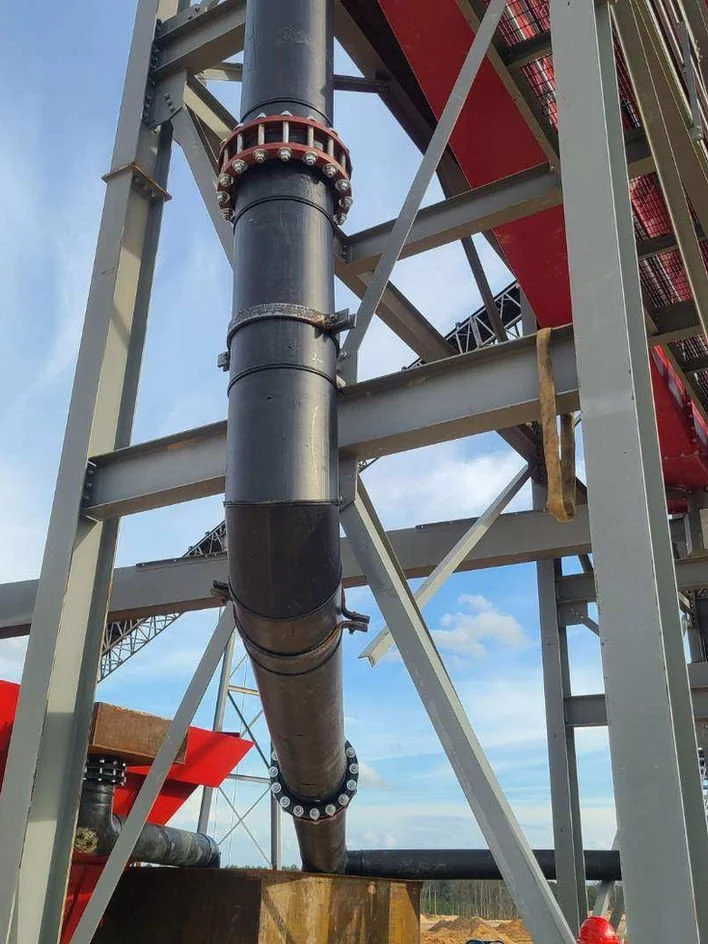 A large industrial pipe supported by a metal framework, part of a construction site with an overcast sky in the background.