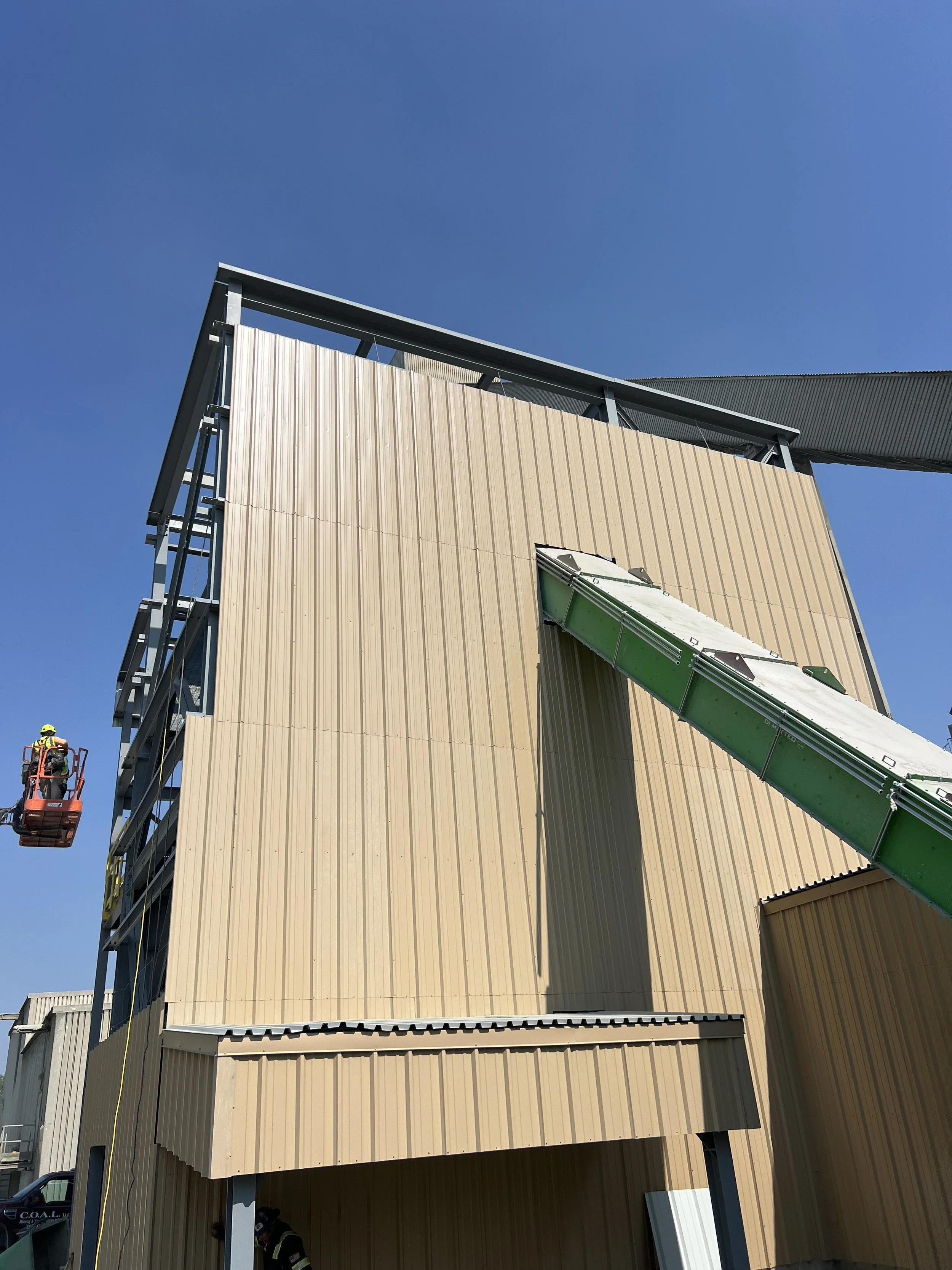 Construction site with a building under construction, featuring beige vertical metal siding, with a green construction chute attached to the side, and worker in a safety vest and helmet visible at the bottom left.
