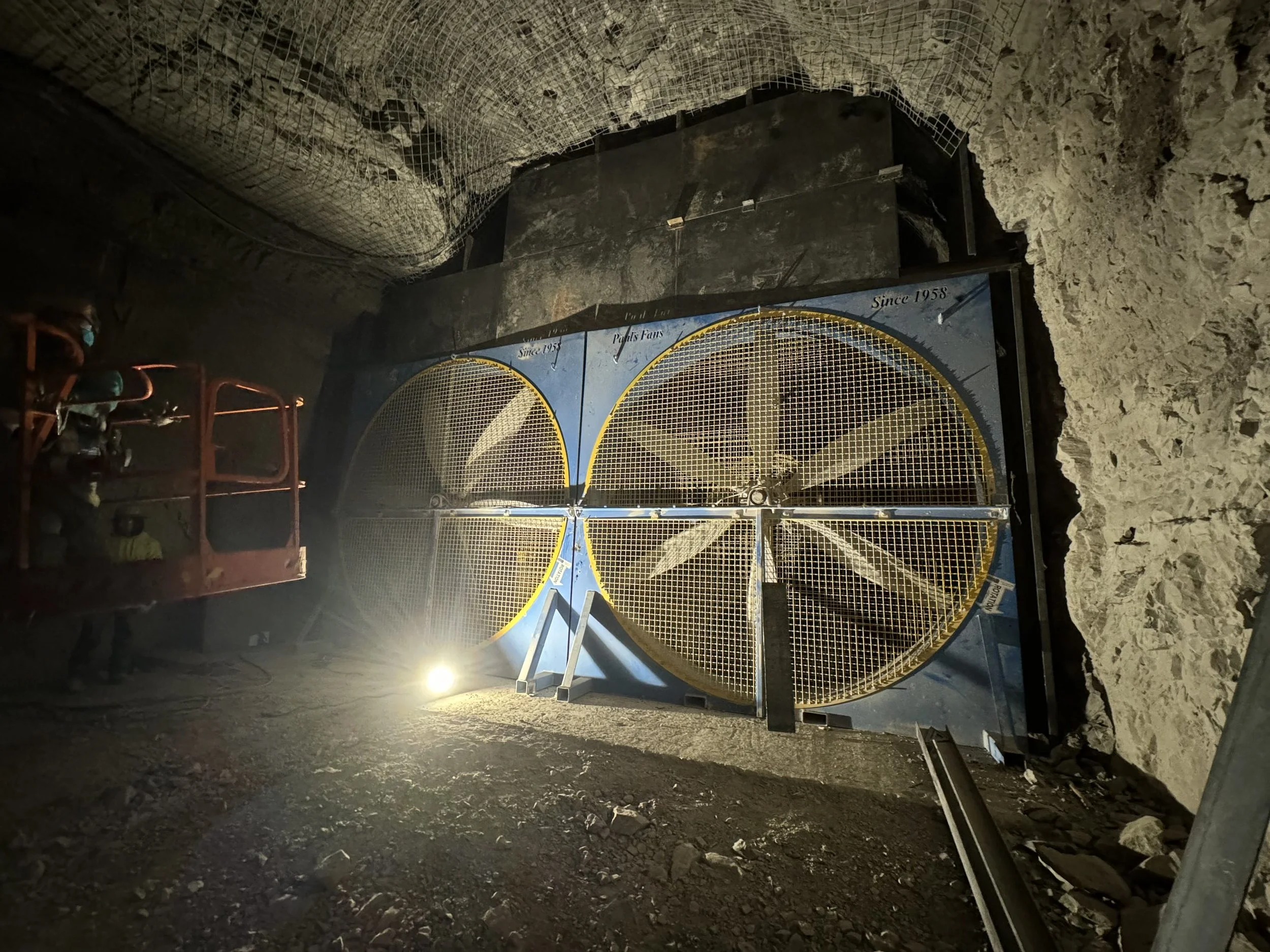 Underground construction site with a large metal tunnel gate, rocky walls, and construction equipment. Illumination from a bright light source on the ground.